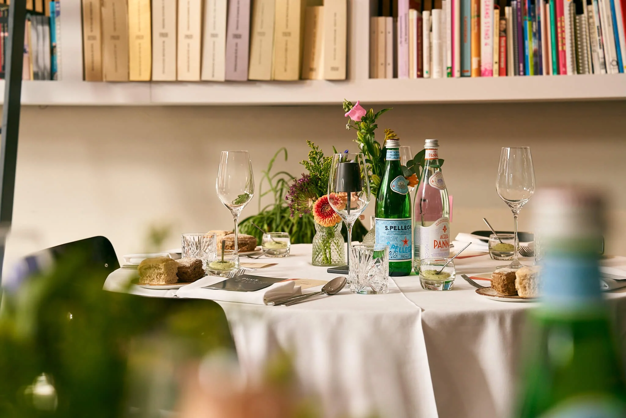 Een gezellige eettafel met drankjes, bloemen en hapjes, op de achtergrond boeken op een muurplank.