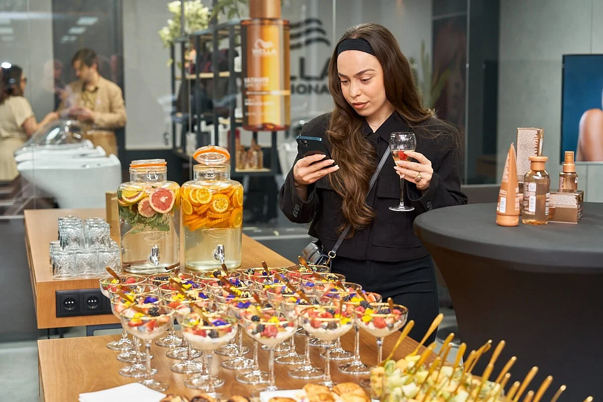 Een vrouwertje met lang, bruin haar, gekleed in zwart, houdt een glas wijn en kijkt op haar smartphone tijdens een feestje of evenement, omgeven door hapjes en drankjes op een tafel.