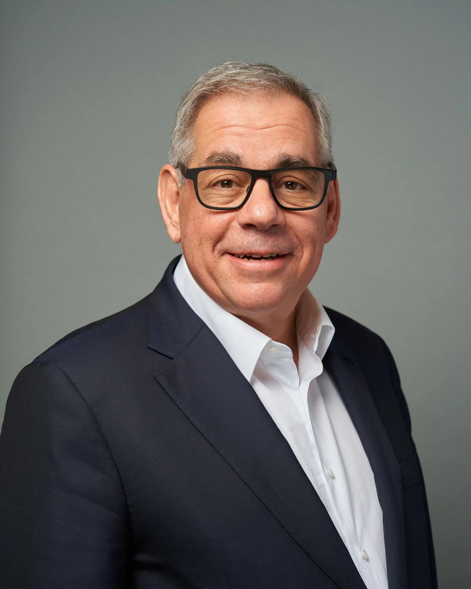 Een man met grijs haar, zwarte bril en een witte overhemd draag een donkerblauw pak, glimlacht naar de camera, tegen een effen grijze achtergrond.
