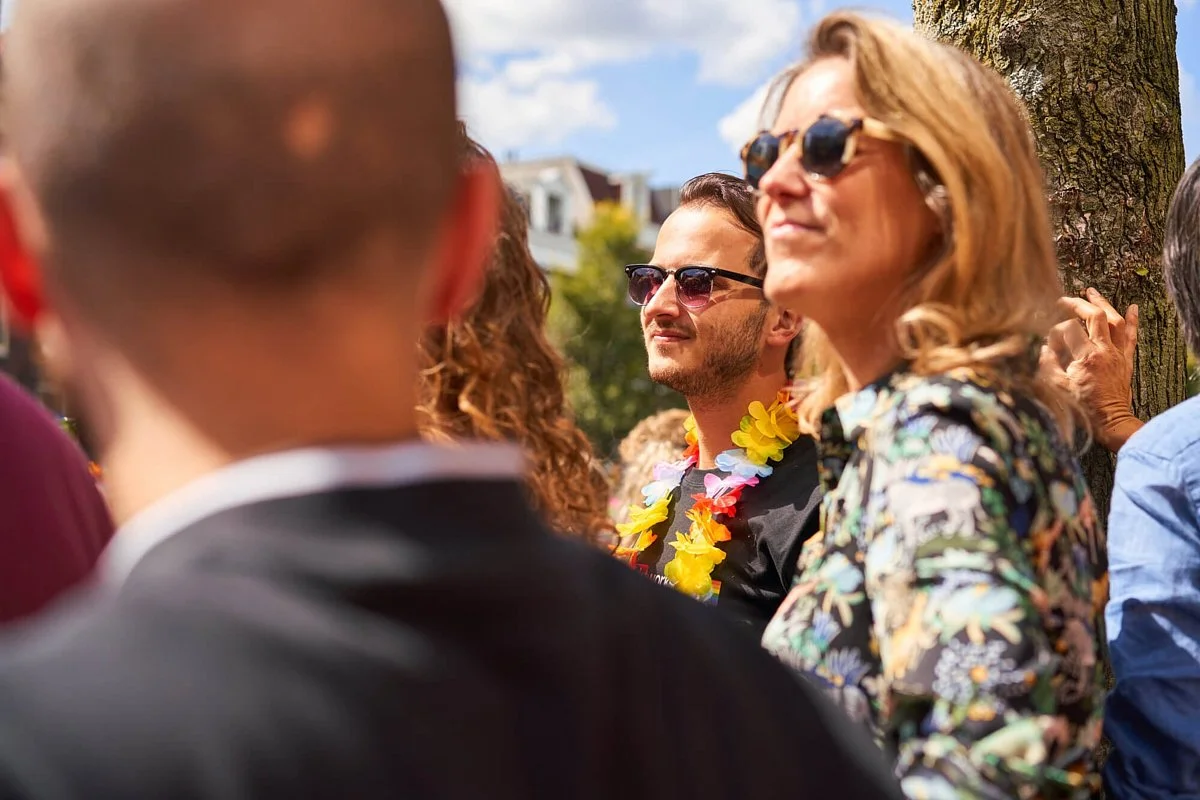 Mensen op een buitenfeestje, gekleed in feestelijke en kleurrijke kleding, met zonnebrillen en bloemenkransen, onder een blauwe hemel met een boom.