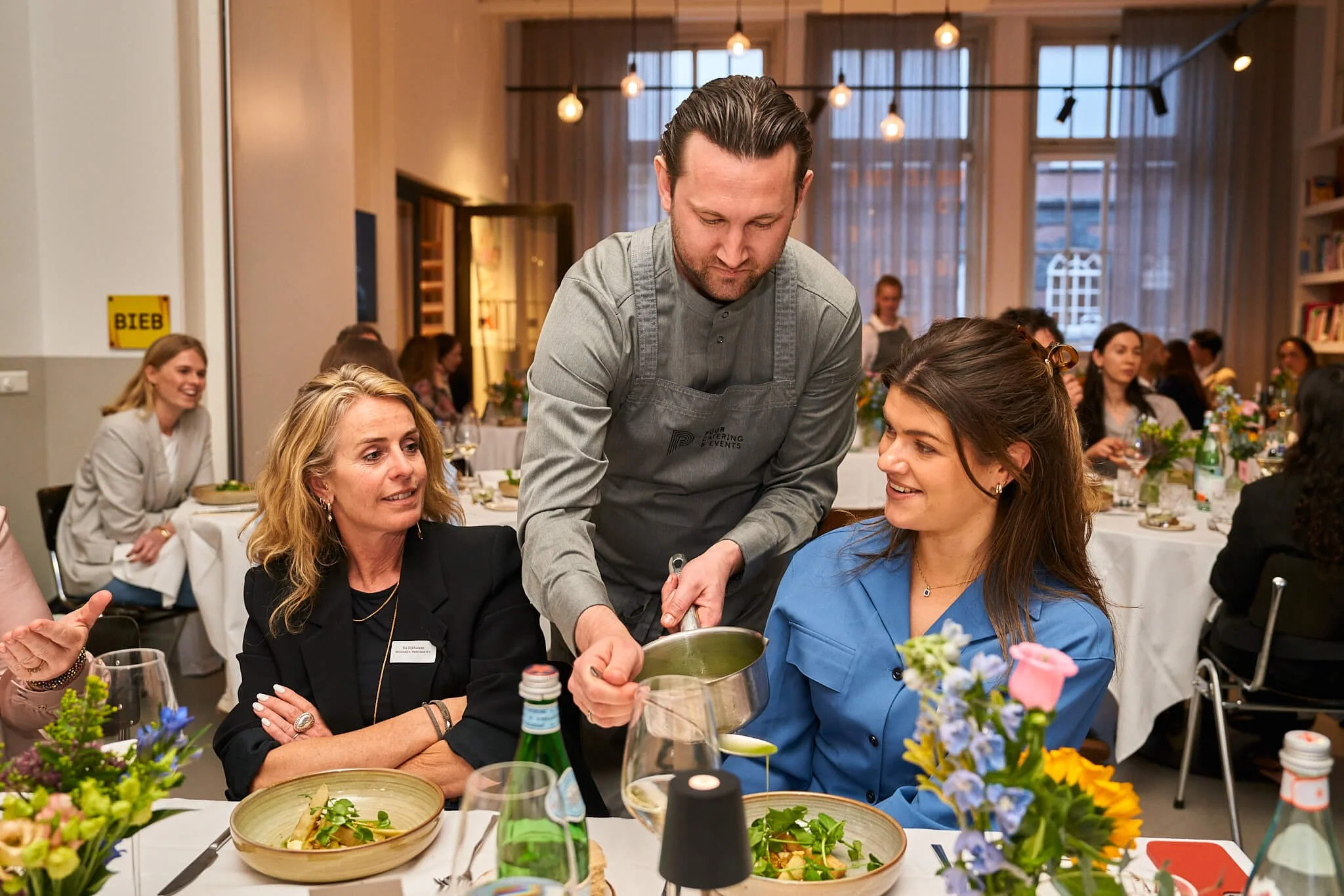 Koks serveert gerecht aan een vrouw tijdens dinersetting met meerdere mensen in een feestzaal.