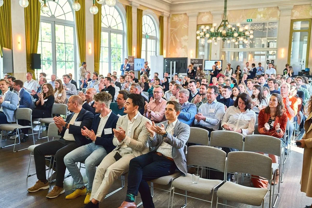 Een grote groep mensen zit in een conferentiezaal, applaudisserend en luisterend naar een presentatie, met grote ramen en een kroonluchter in het plafond.