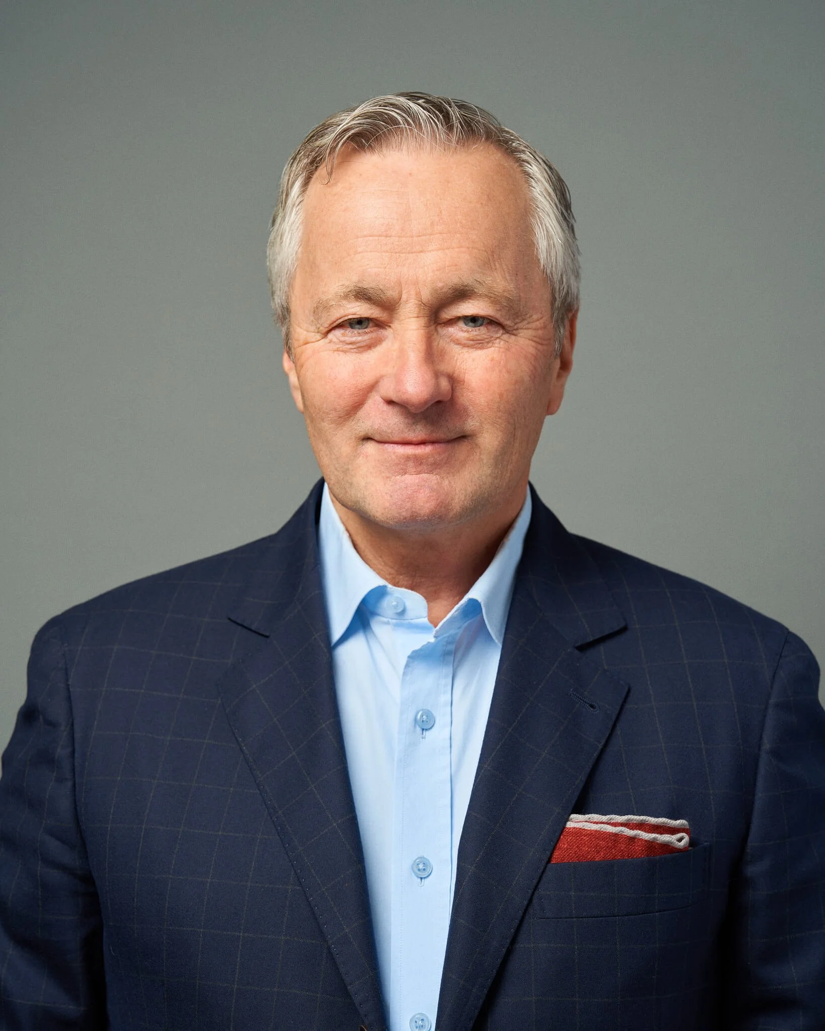 Portret van een volwassen man met grijs haar, glimlachend, gekleed in een donkerblauw blazer en een lichtblauwe overhemd, tegen een effen grijze achtergrond.