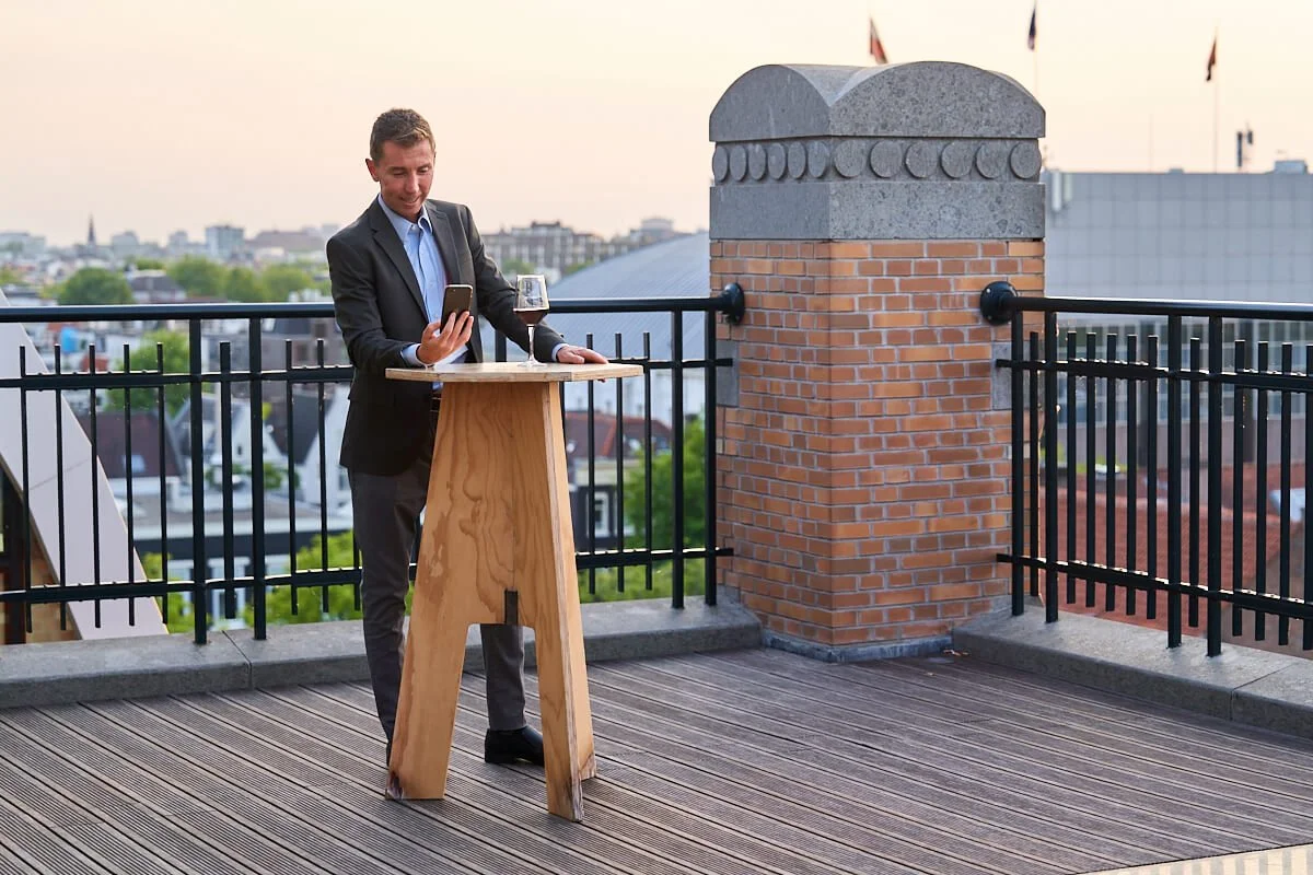 Man in pak met wijnglas op dakterras, kijkend naar zijn telefoon