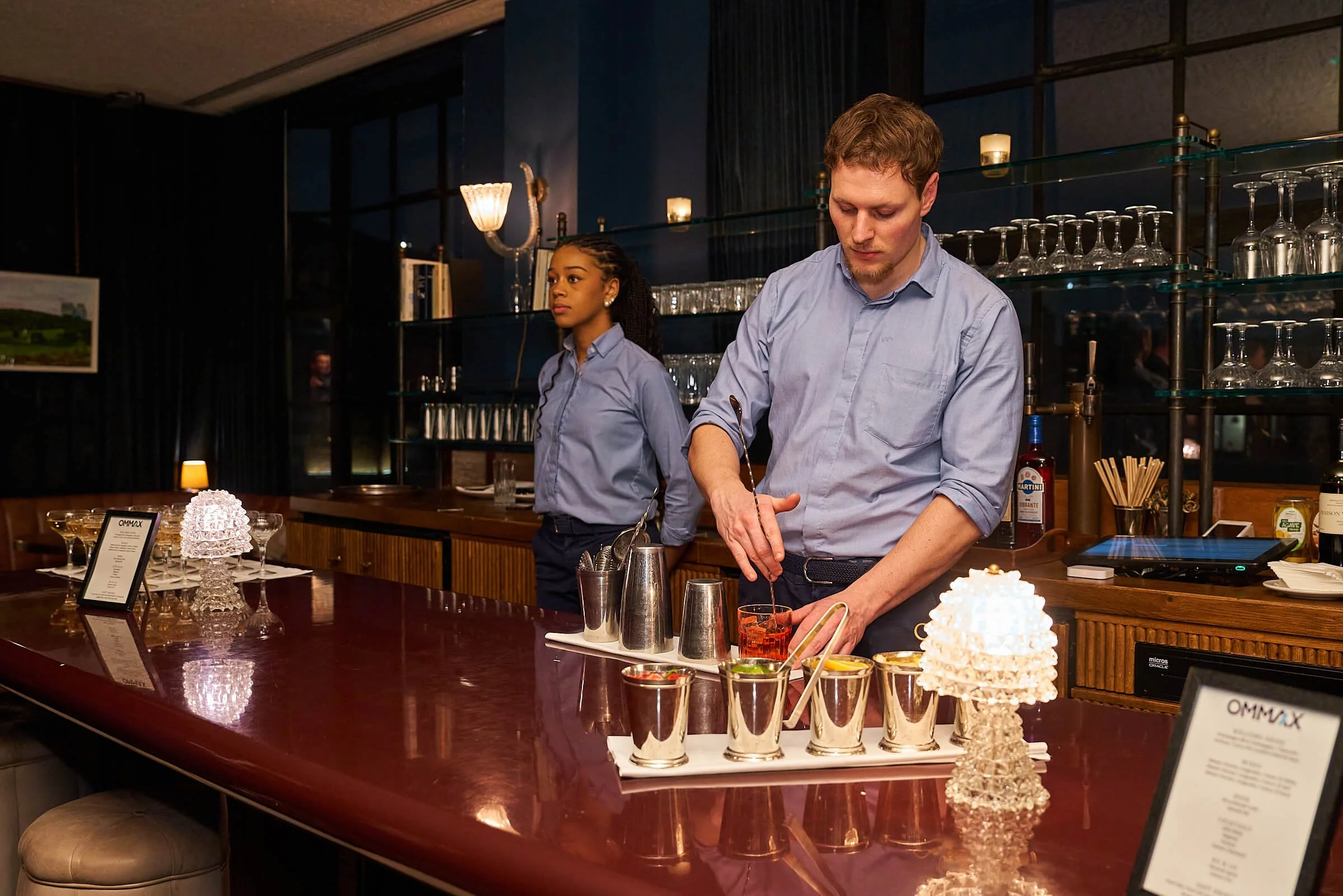 Bartender bereidt cocktails in een bar, met een vrouwelijke assistent op de achtergrond, in een gezellig, sfeervol interieur.