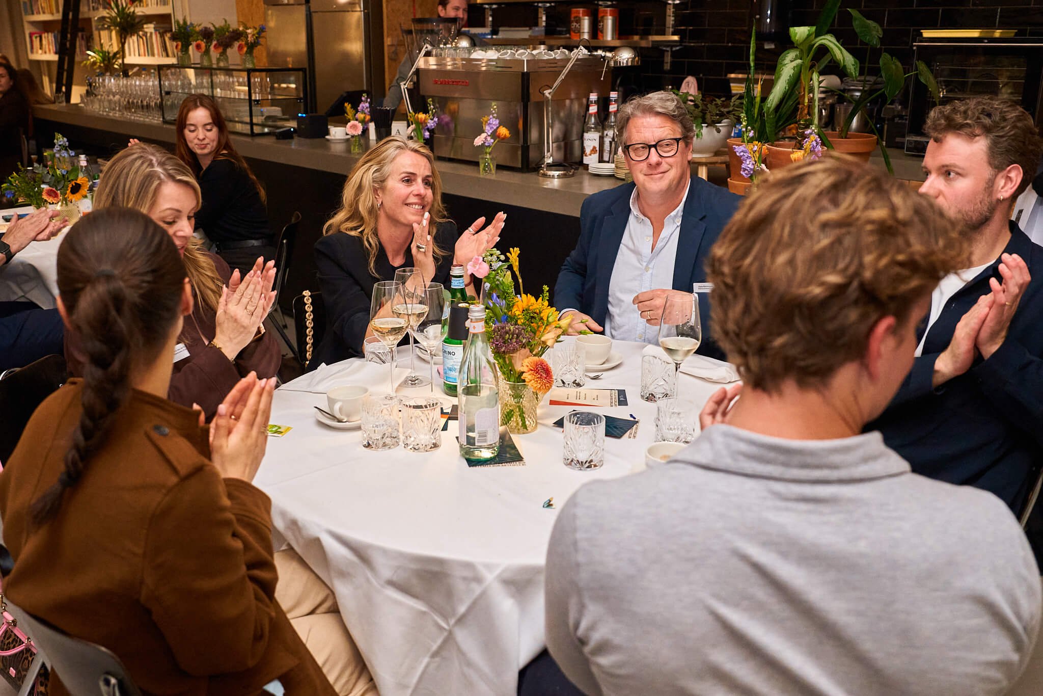 Een groep mensen zit aan een ronde tafel in een restaurant of op een evenement, applaudisseert en kijkt naar iemand die niet in het beeld zichtbaar is, met bloemen en drankjes op de tafel.