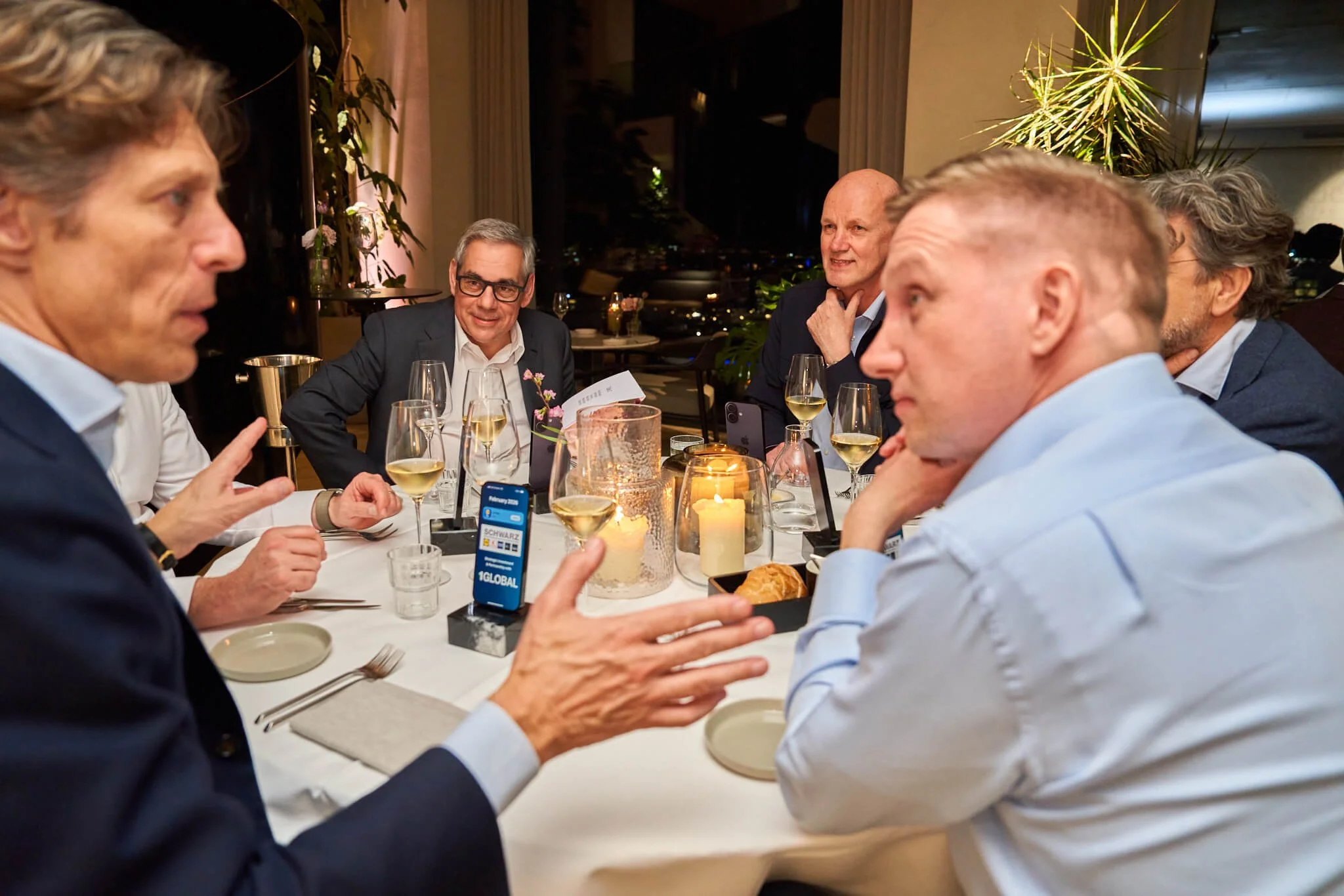 Vijf mannen die in een restaurant of een formeel diner zitten aan een ronde tafel met glazen witte wijn, kaarsen en een gebakje. Ze lijken discussiëren of praten met elkaar.