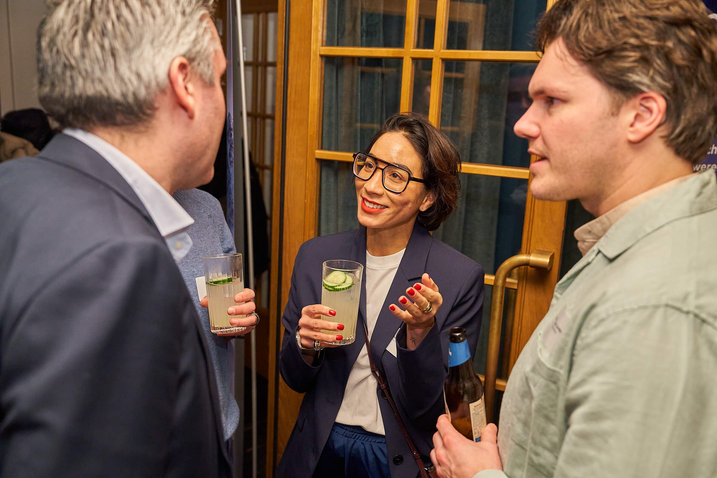 Drie mensen die communiceren tijdens een sociale bijeenkomst, twee mannen en één vrouw, met glazen drinken in de hand, lachen en praten in een gezellige binnenruimte.