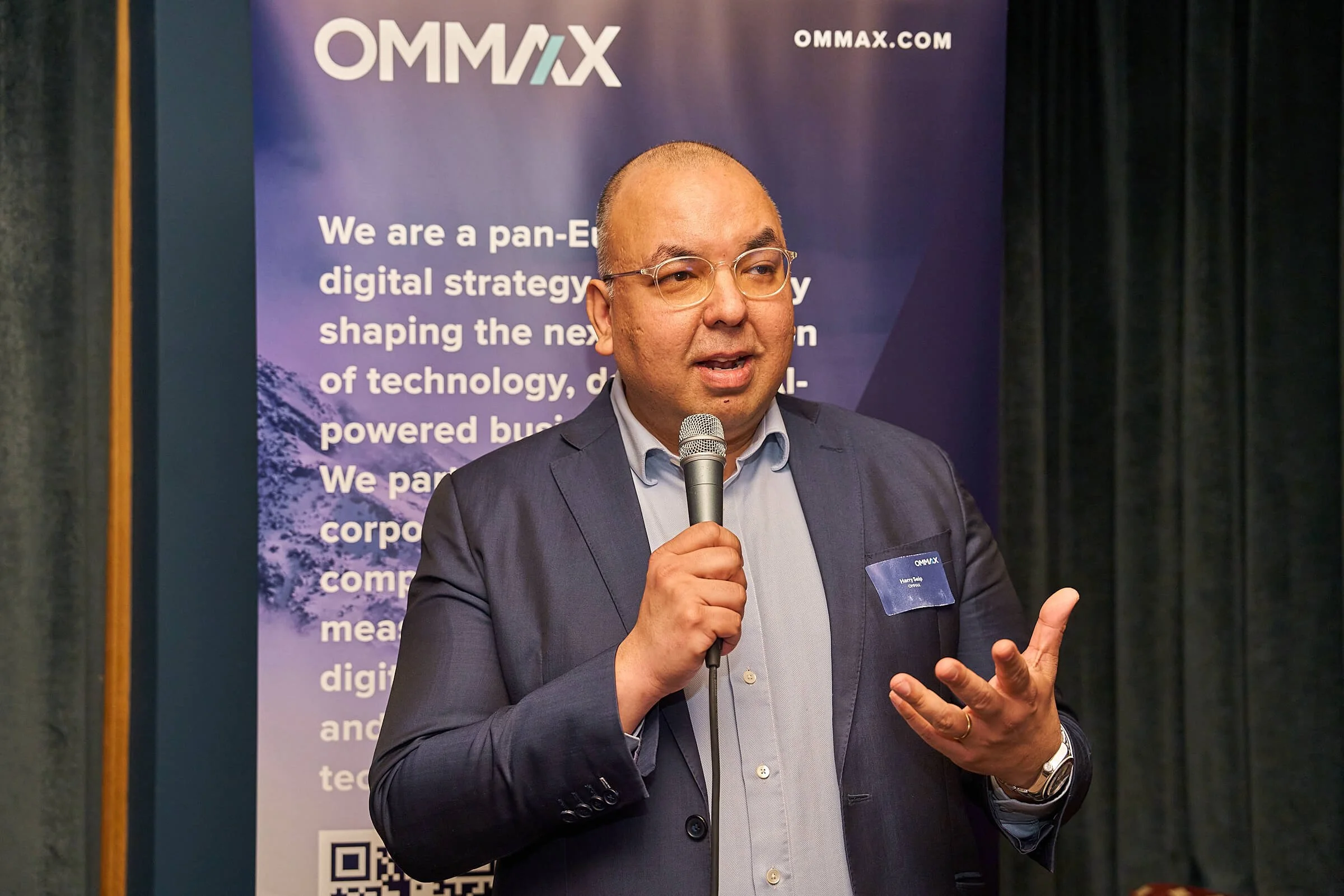 Man in zakelijke kleding geeft een presentatie met microfoon tijdens een evenement, met een OMMAX-banner op de achtergrond.