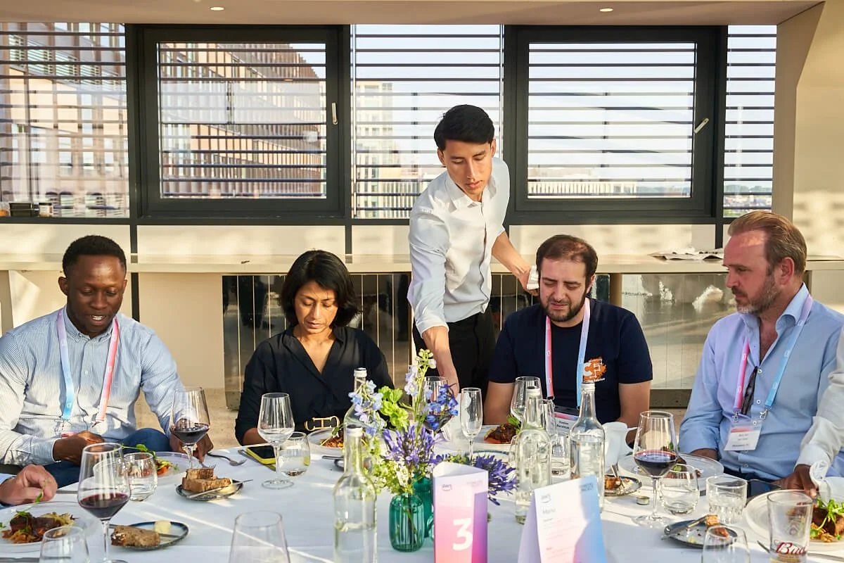 Mensen zitten aan een tafel met eten en wijn, een ober serveert eten, op een zonnig terras met grote ramen en uitzicht op de stad.