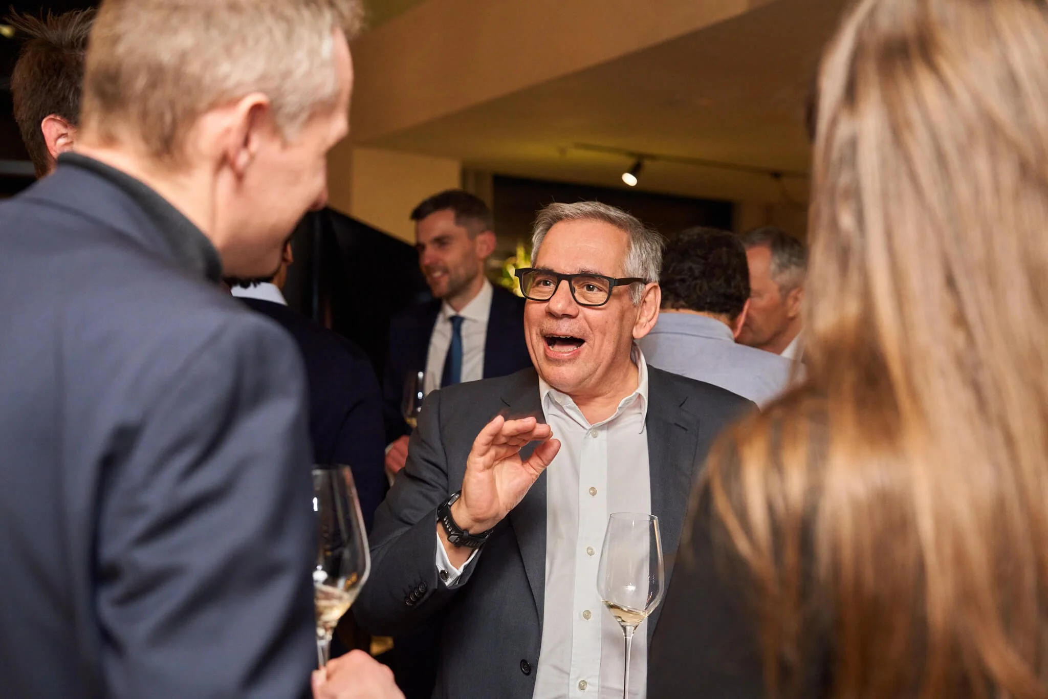 Man in grijs pak en witte blouse die spreekt tijdens een netwerkevenement met wijnglazen in de hand, omringd door andere mensen in gesprek.