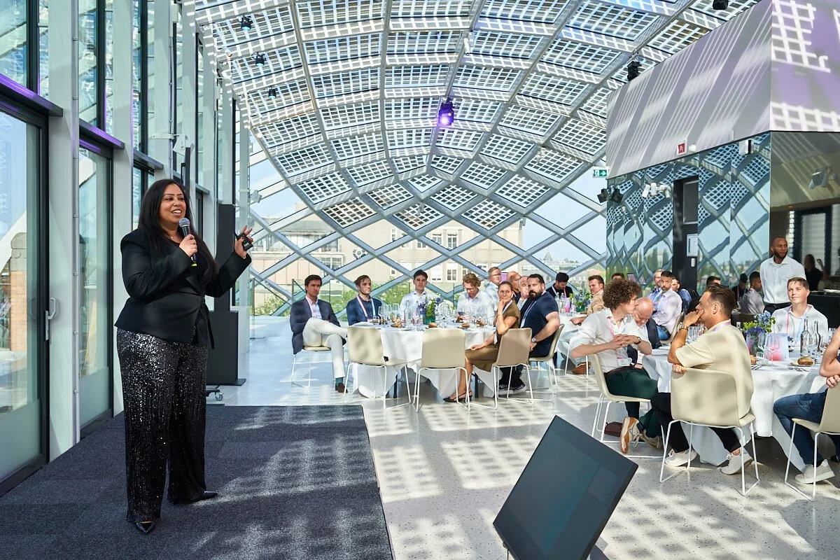 Een vrouw houdt een presentatie met een microfoon voor een groep mensen die aan tafels zitten in een moderne, glazen constructie. De zon schijnt door het gebogen, glasgevels.