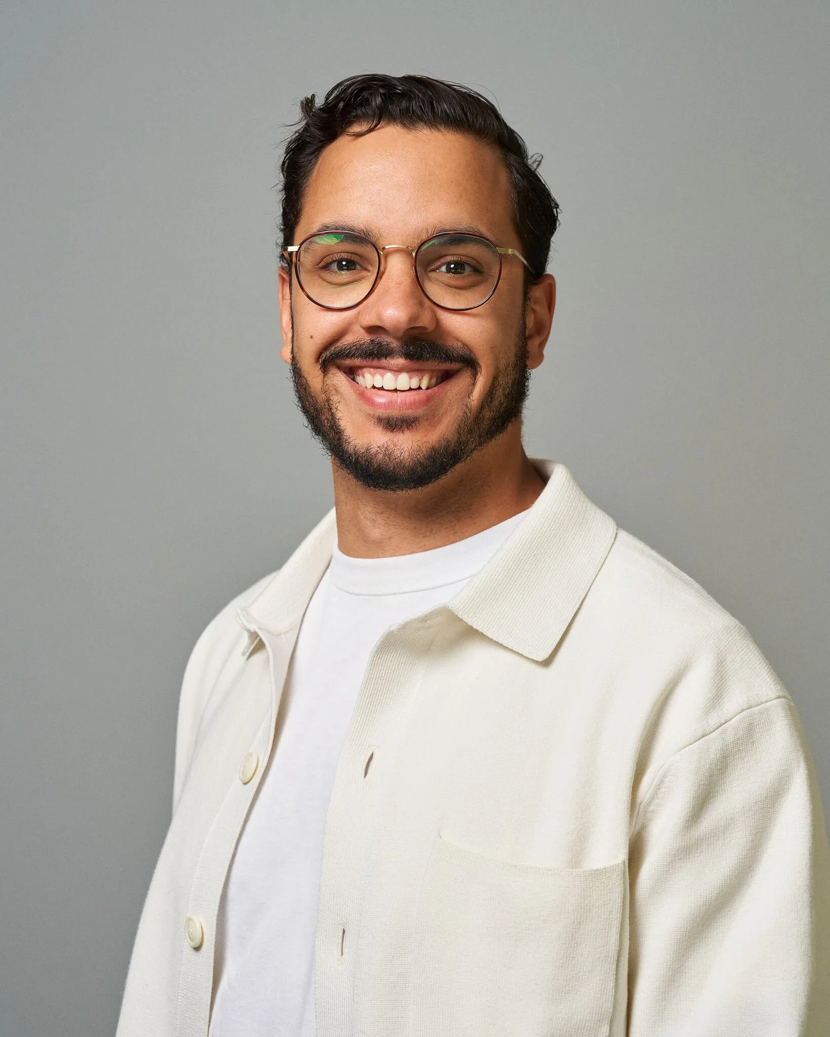 Man met bril en baard, lachend, dragen een witte trui op een grijze achtergrond.