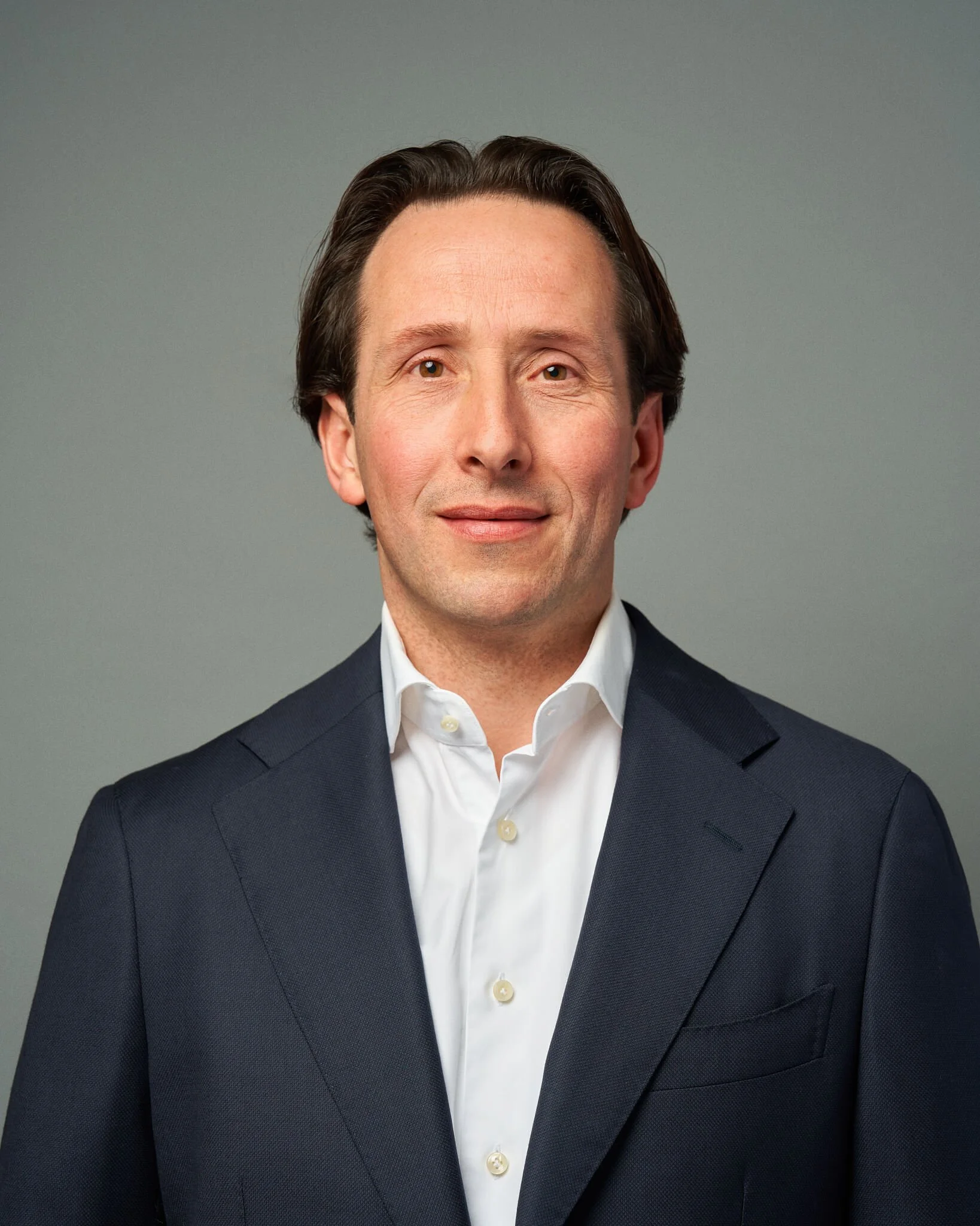 Een man met donker haar gekleed in een wit overhemd en een donkerblauwe jas, glimlachend voor een grijze achtergrond.