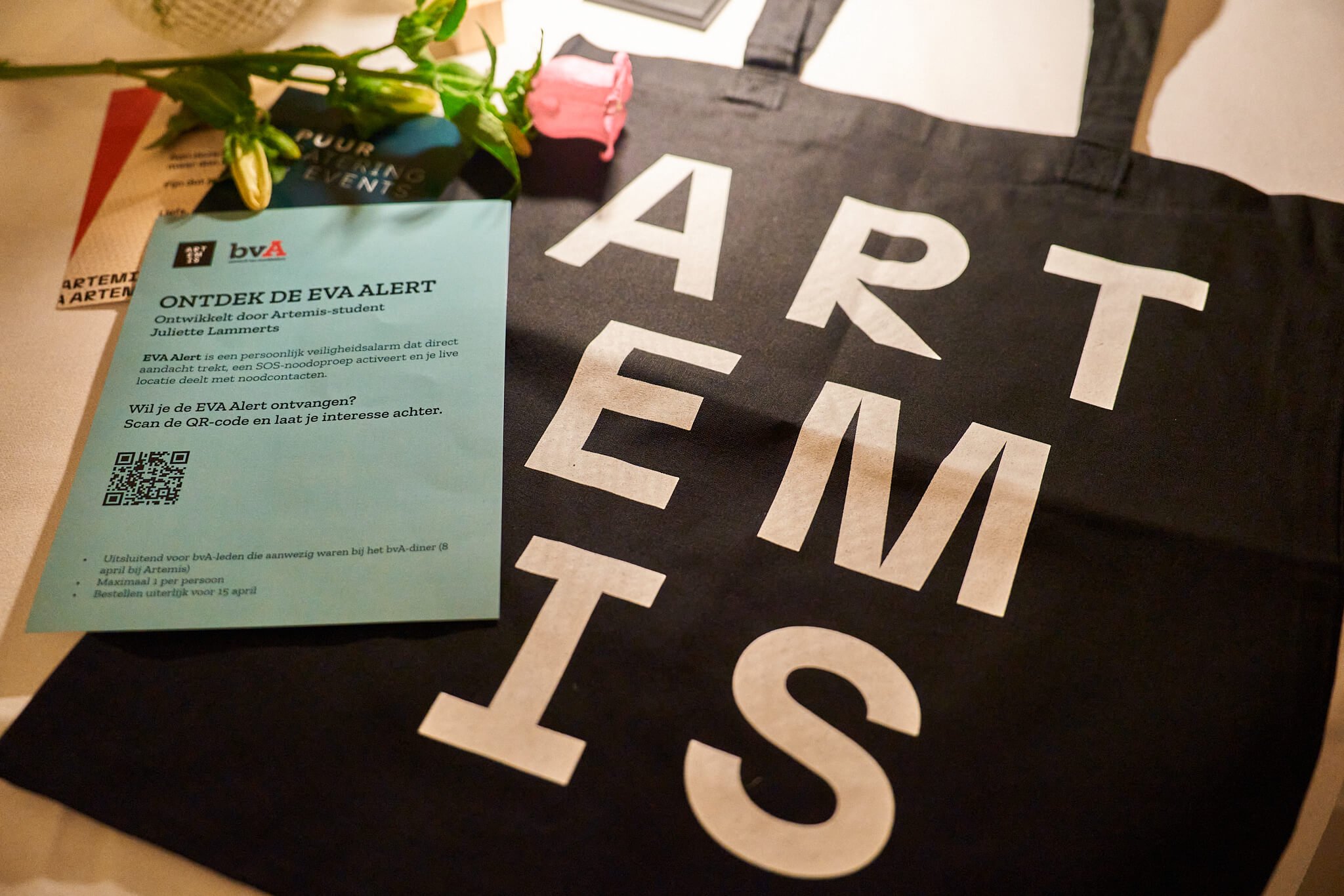 Een zwarte tas met witte letters waarop 'ART EN W' staat, een stuk papier met tekst en een QR-code, een roze bloem, en een paar witte bloemen op een tafel.