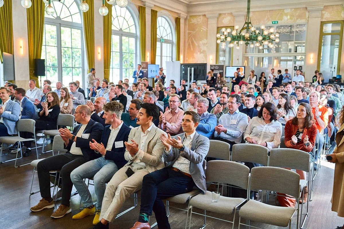 Een grote groep mensen zit in een zaal en applaudisseert tijdens een conferentie of bijeenkomst, met grote ramen en groene gordijnen op de achtergrond.