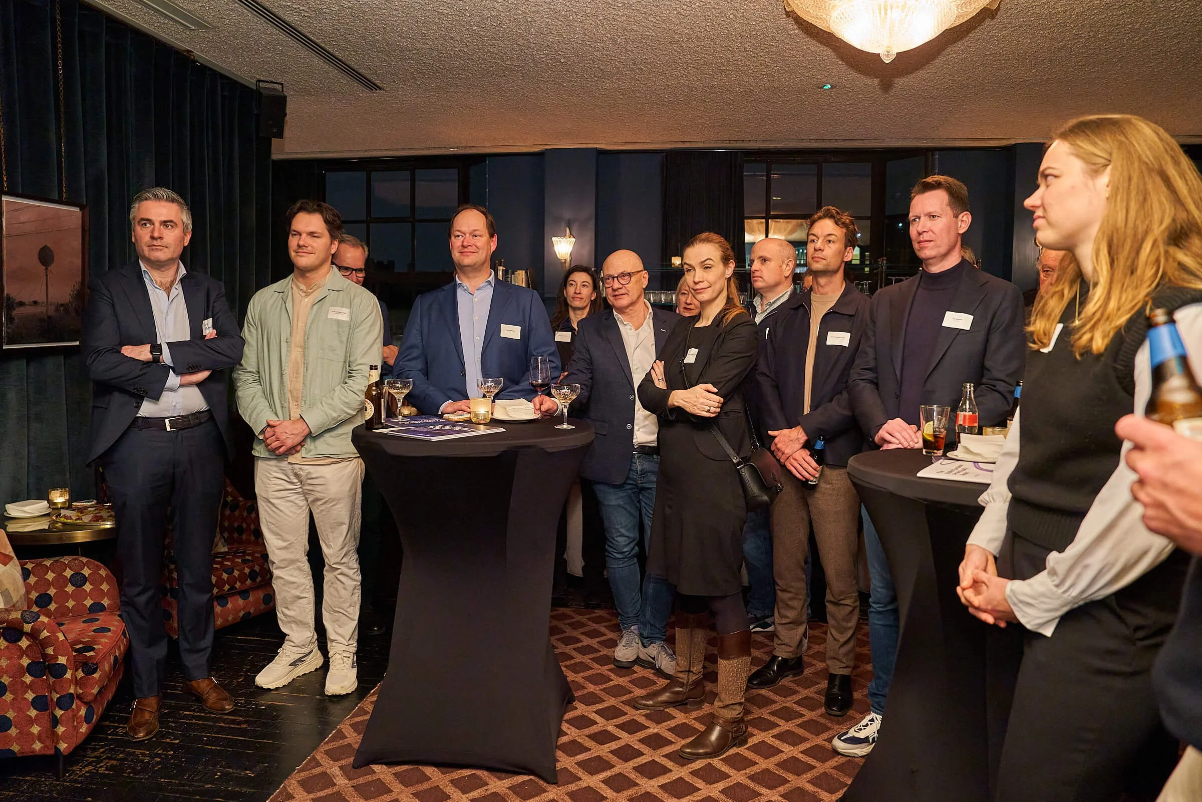 Mensen staan in groep tijdens een netwerkevenement in een restaurant of bar, met high tables, drankjes en snacks, terwijl ze luisteren naar iemand die spreekt.