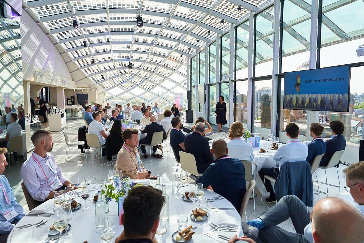 Een grote groep mensen zit aan ronde tafels in een lichte, moderne zaal met veel glas en een gebogen plafond, tijdens een zakelijke of conferentie-evenement.