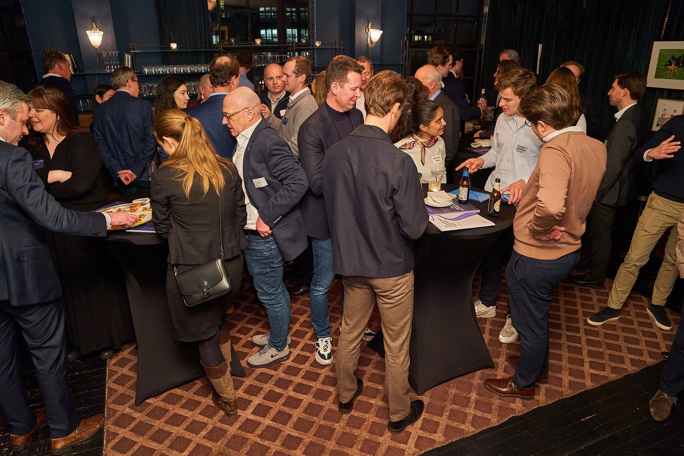 Een groep mensen bij een netwerkborrel of evenement in een binnenruimte met lage verlichting, waar ze staan rond hoge ronde tafels, en drankjes en informatiemateriaal op tafel liggen.