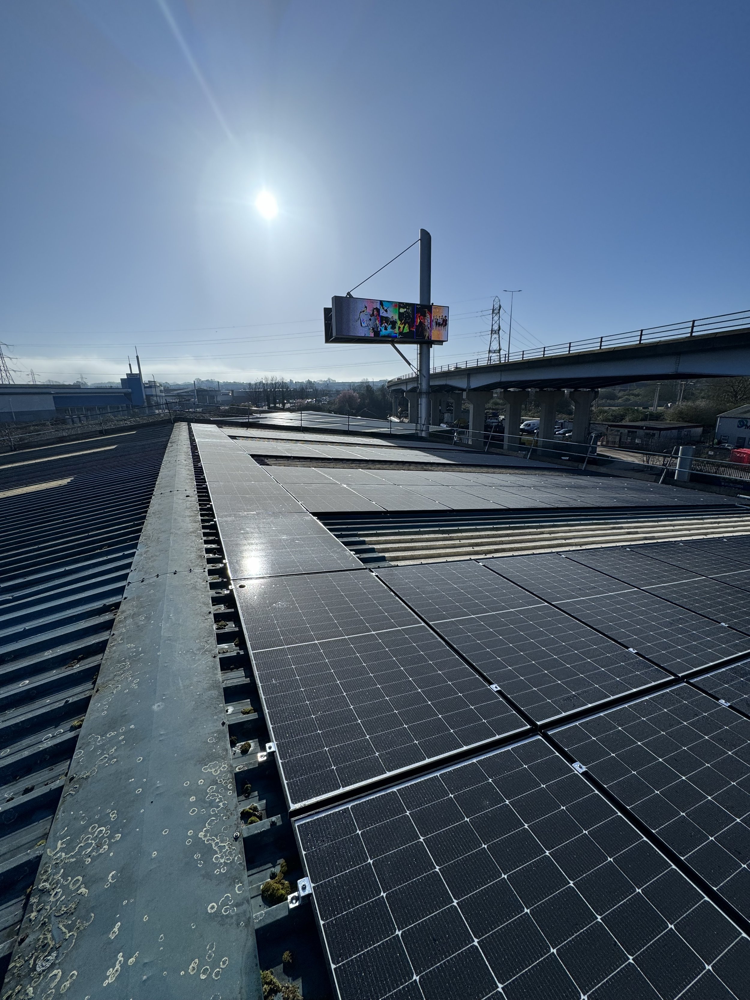 Solar panels installed on a roof under a sunny sky, with an electronic billboard and highway in the background.