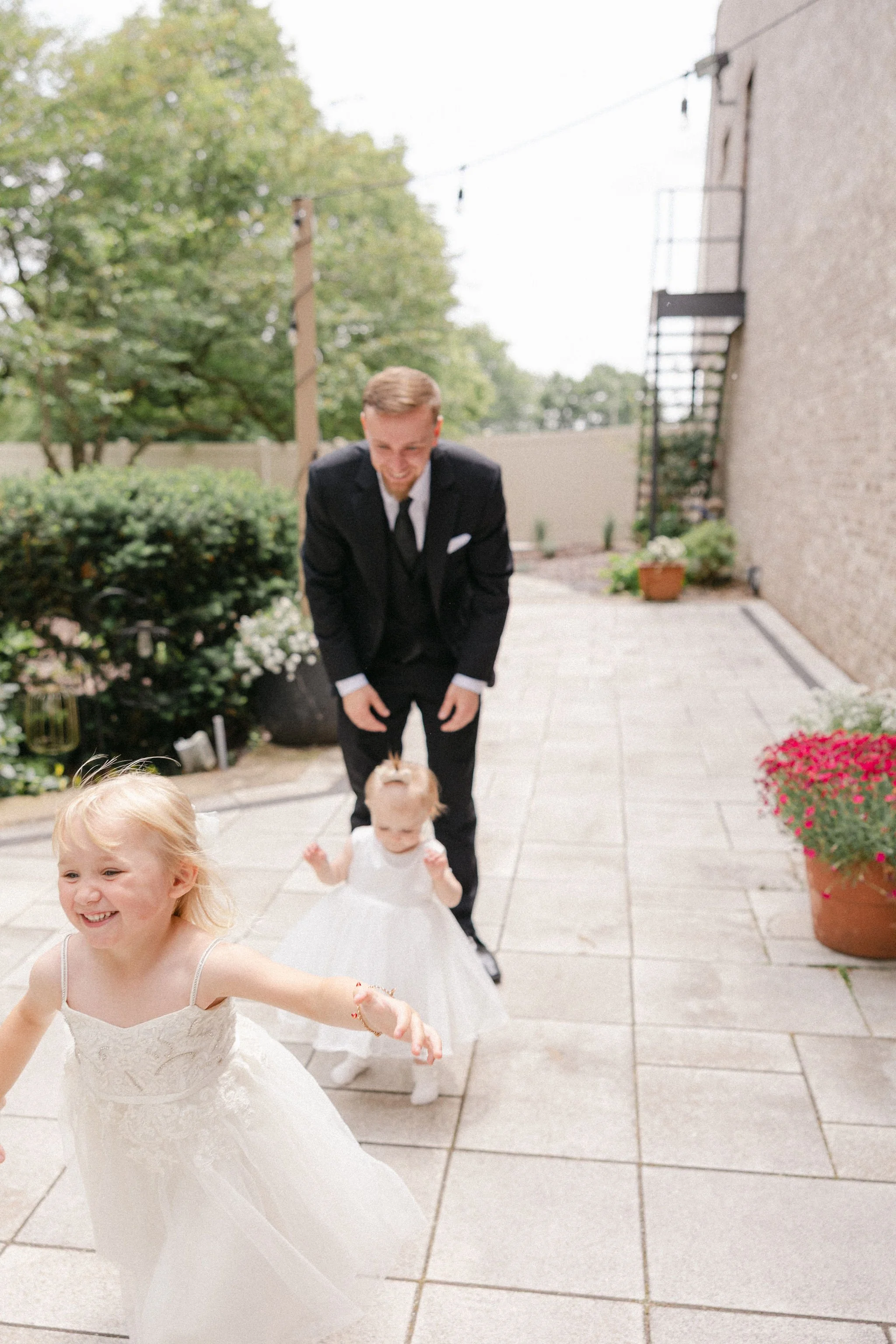 A man in a black suit and two young girls in white dresses playing or dancing outdoors on a tiled patio, with potted flowers and greenery in the background.