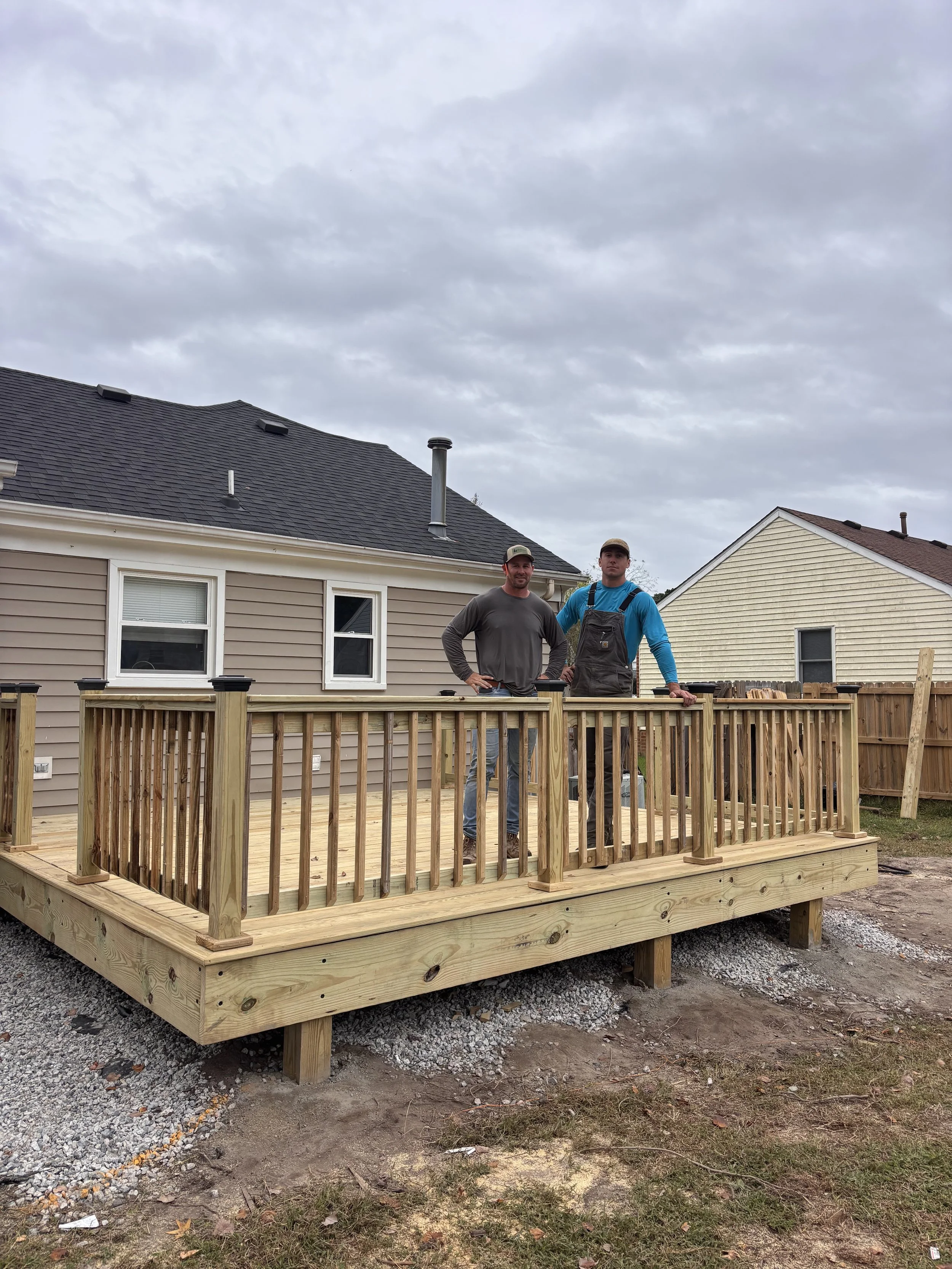 Two men stand on a newly built wooden deck in a backyard, with houses and cloudy sky in the background.