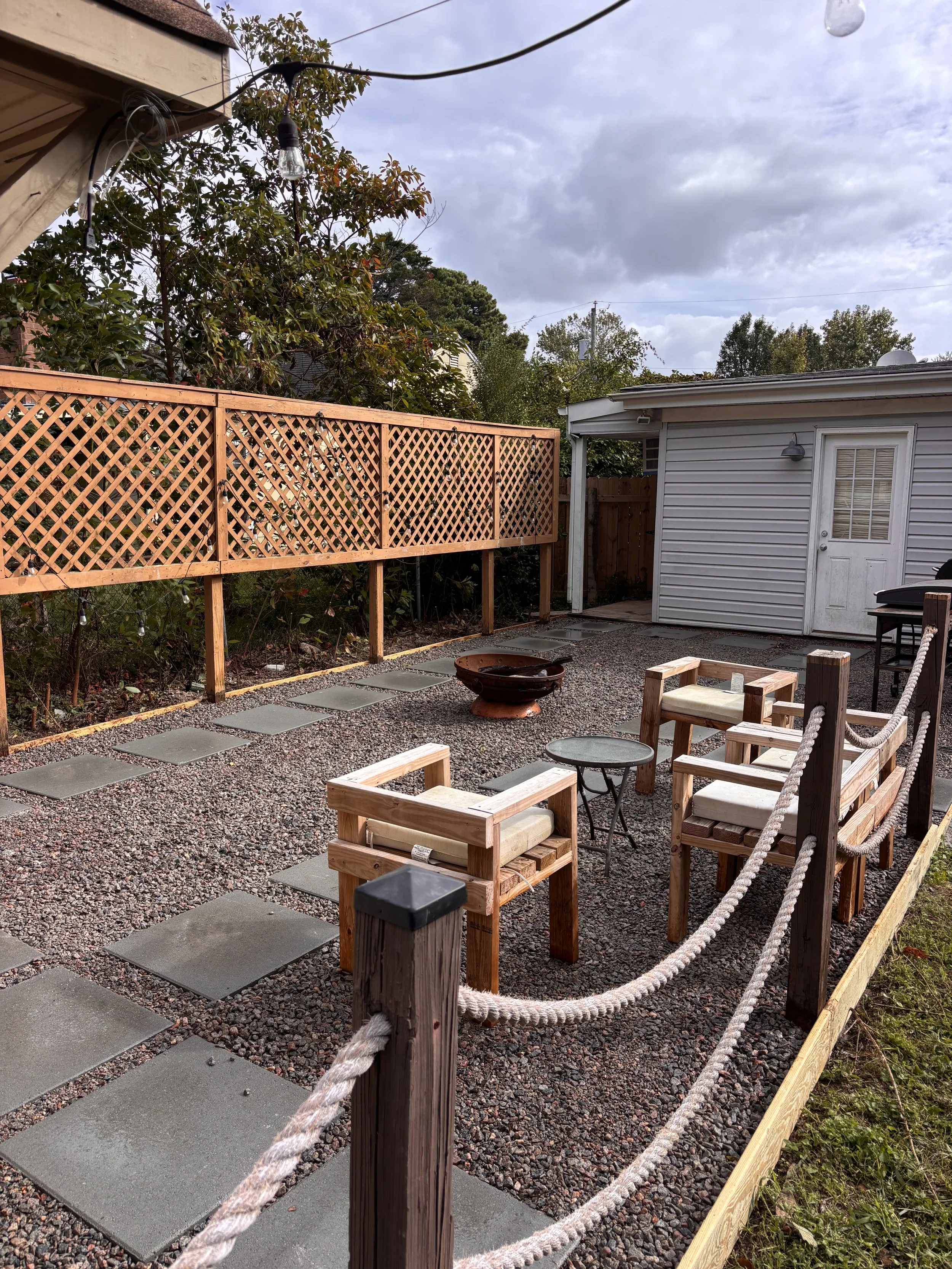 A backyard patio with wooden patio furniture, a small side table, a fire pit, gravel ground, and a wooden fence, with a gray building in the background and cloudy sky above.