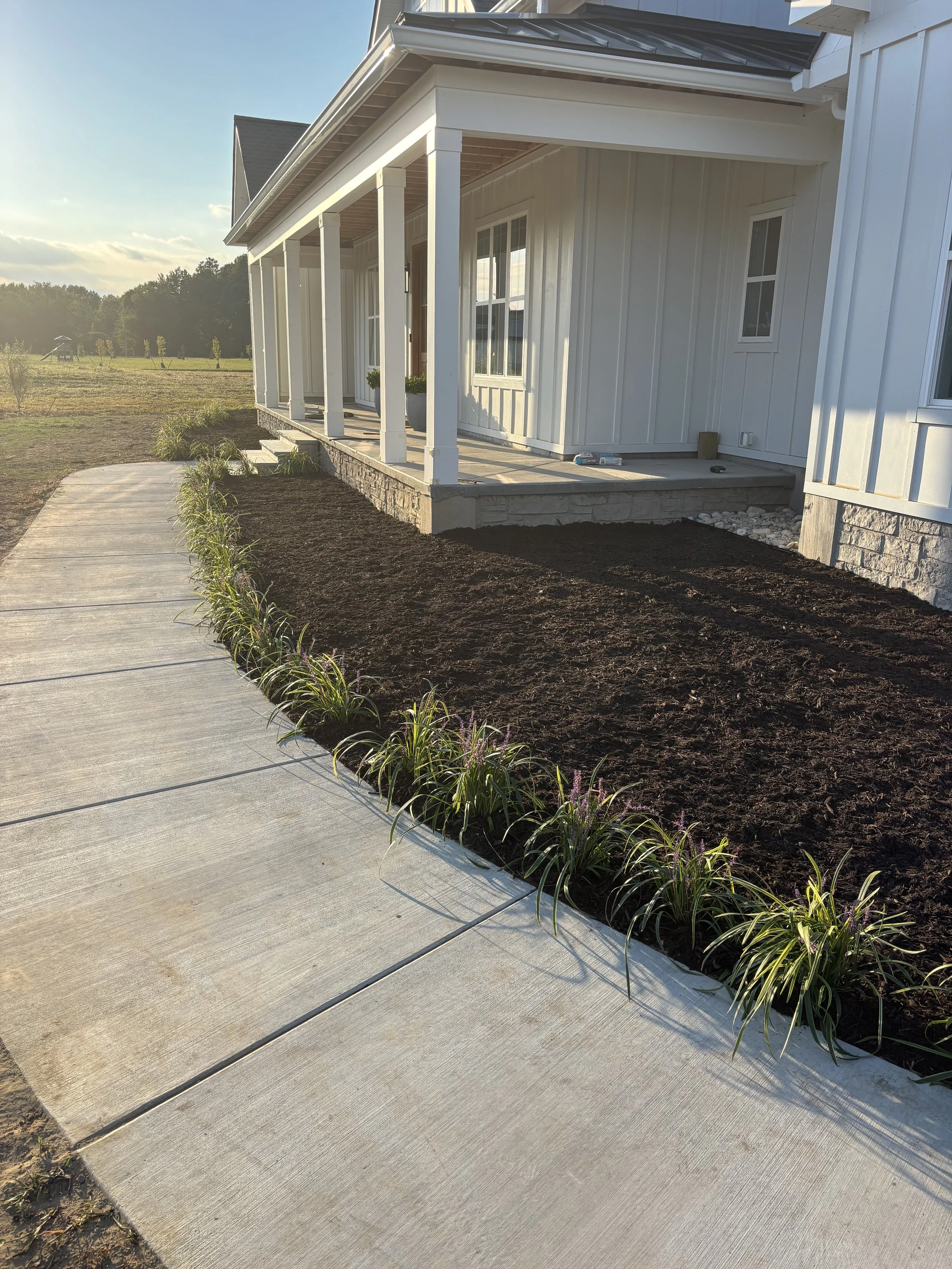 A two-story brick house with a green roof and arched window above the front door decorated with a holiday wreath. The house has four windows and outdoor lanterns flanking the door. There is well-maintained lawn and landscaped garden with shrubs and a rock-bordered flower bed. Tall leafless trees are in the background, indicating winter.