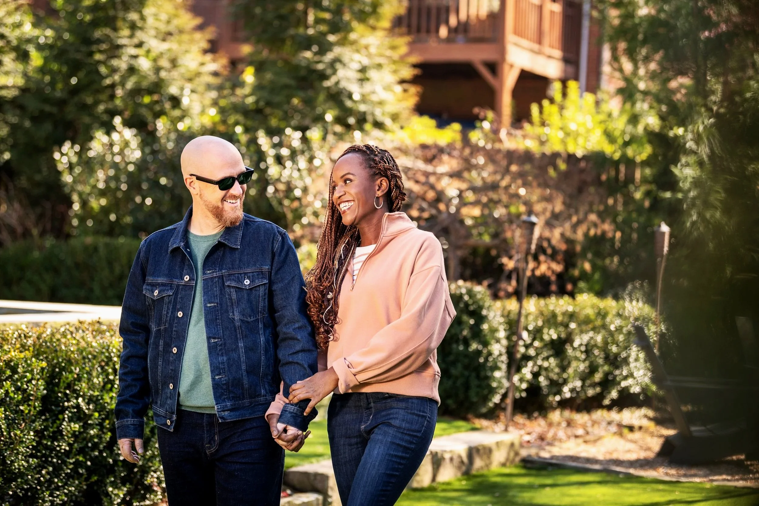 Echo Frames by Amazon. Happy couple walking outdoors in a park, holding hands and smiling at each other, with lush green bushes and trees in the background on a sunny day.