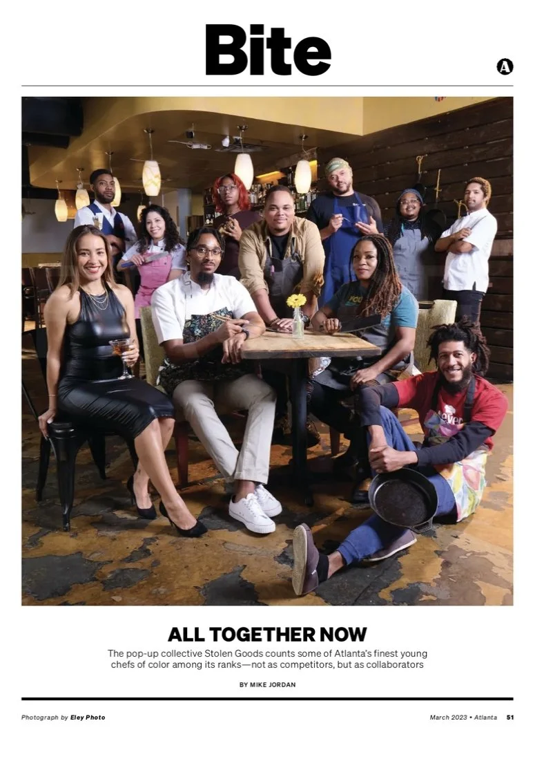 Group of diverse young chefs and restaurant staff gathered together inside a restaurant, posing for a photo, with some seated at a table and others standing behind.