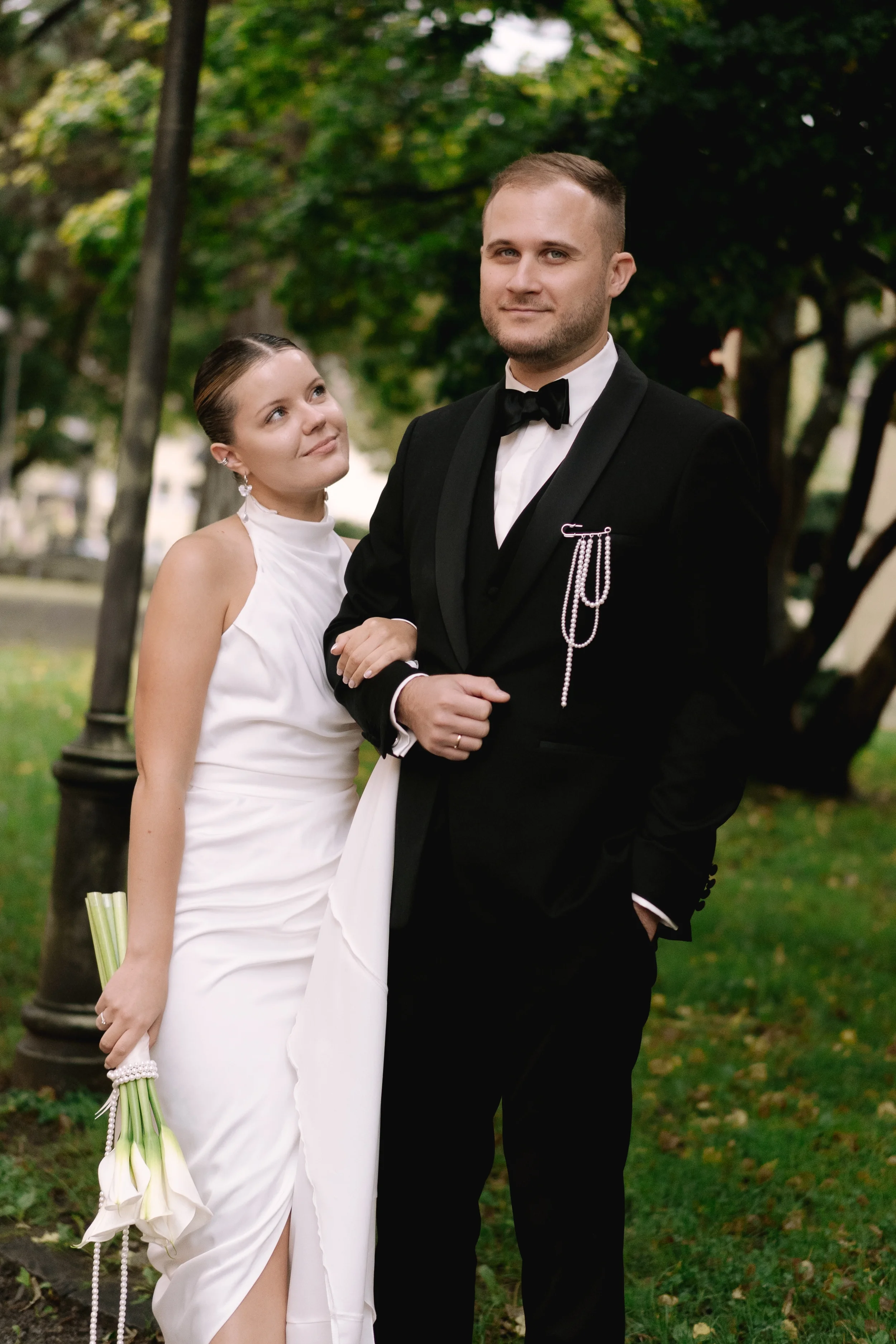 Ein Brautpaar beim Gehen im Park, die Frau trägt ein weißes Brautkleid und hält einen Blumenstrauß, der Mann trägt einen schwarzen Anzug mit Fliege. Im Hintergrund sind Bäume und eine Laterne.