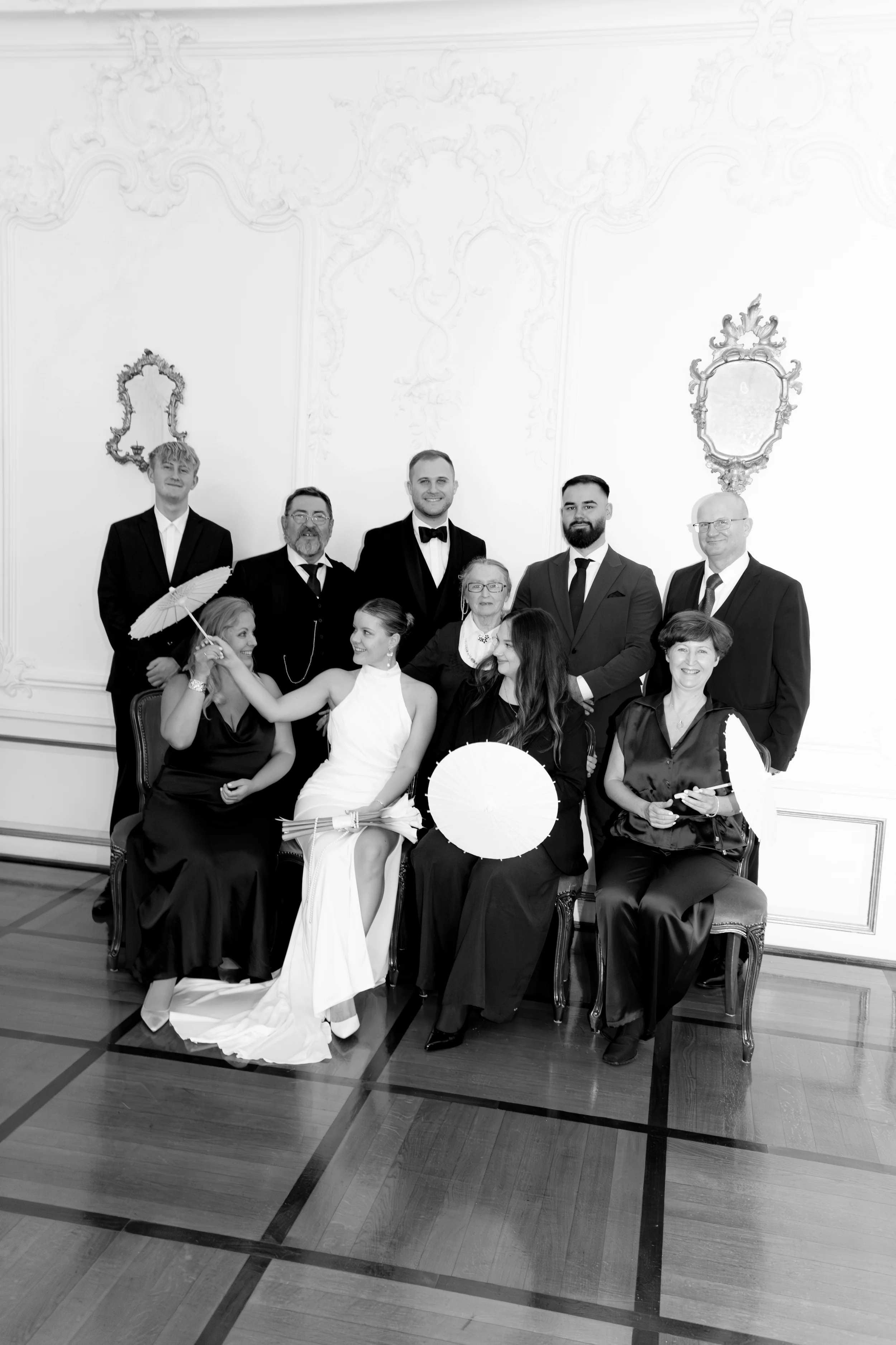 Eine Gruppe von Menschen bei einer formellen Veranstaltung in einem eleganten Raum. Einige sitzen, andere stehen, alle in formeller Kleidung. Es gibt Damen mit weißen und schwarzen Kleidern und Herren in Anzügen, einige mit Krawatten. Zwei Damen halt