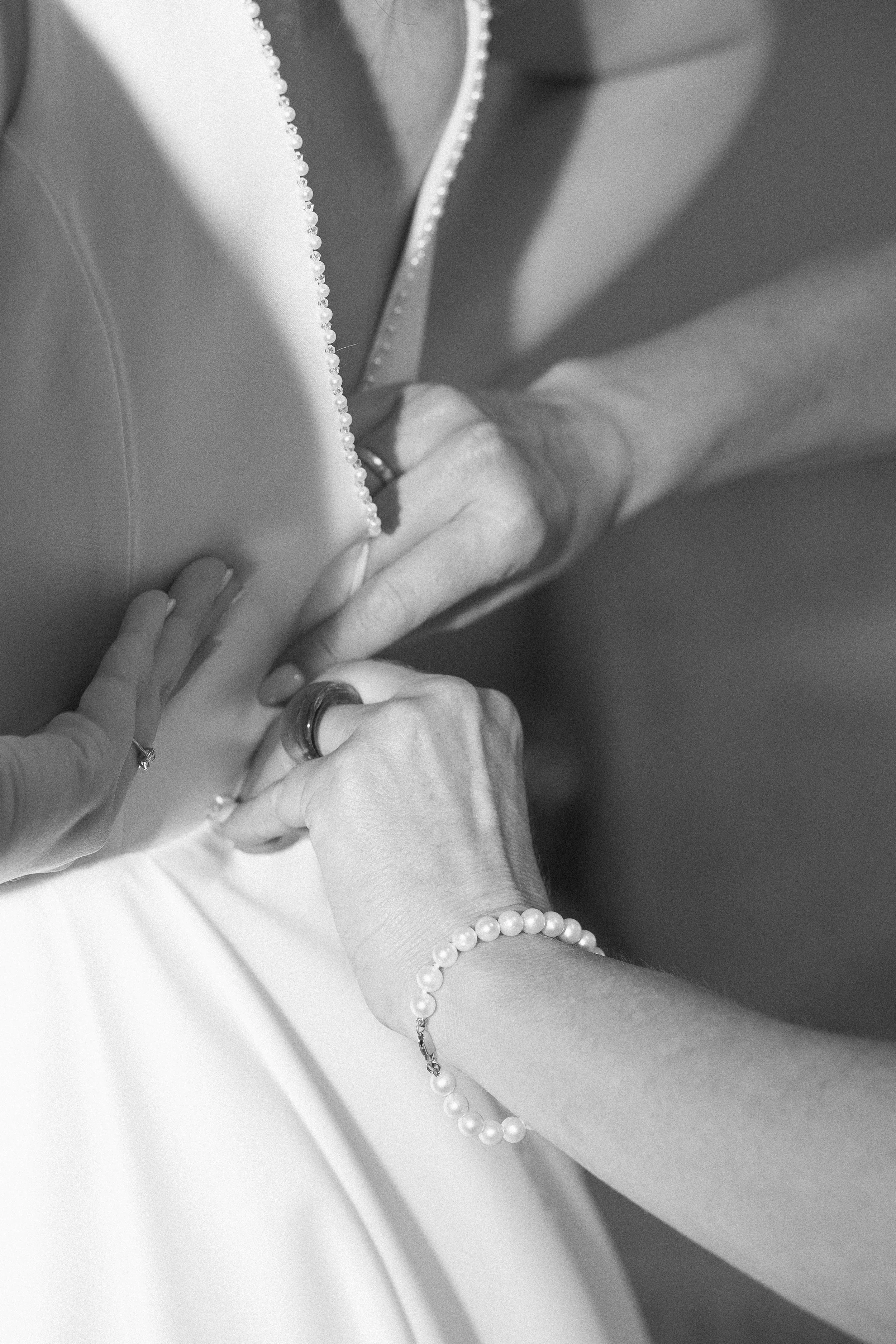 Schwarz-weiß-Foto einer Person, die ein Kleid mit Perlen am Hals trägt. Mehrere Hände greifen nach dem Kleid, eine Hand trägt eine Perlenarmband.