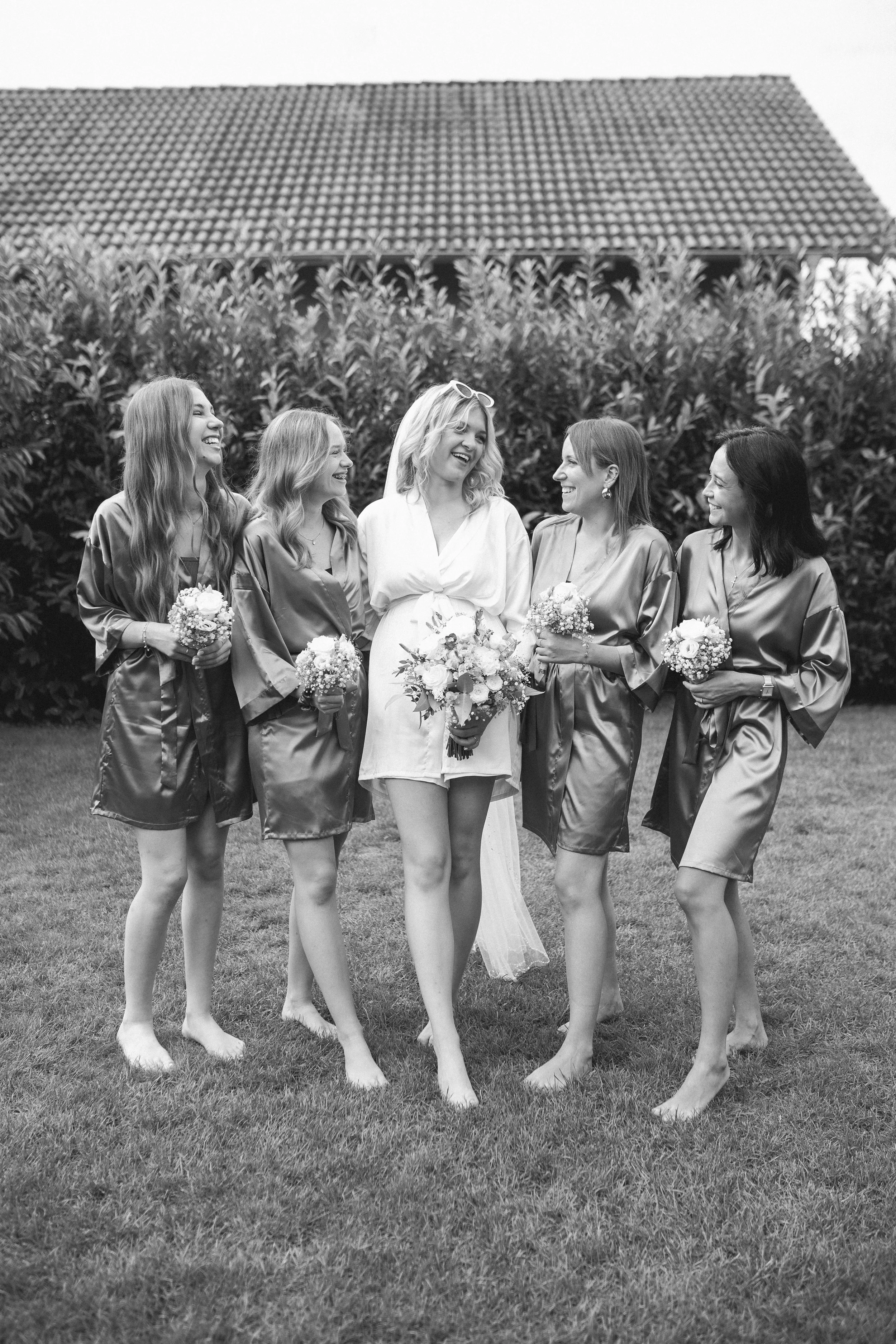 Eine Gruppe von sechs Frauen, darunter eine Braut, die barfuß auf dem Rasen stehen und lachen. Sie trägt ein weißes Hochzeitskleid, während die anderen Frauen rosa Satin-Nachthemden und Blumensträuße halten. Es sieht nach einer Hochzeitsfeier aus.