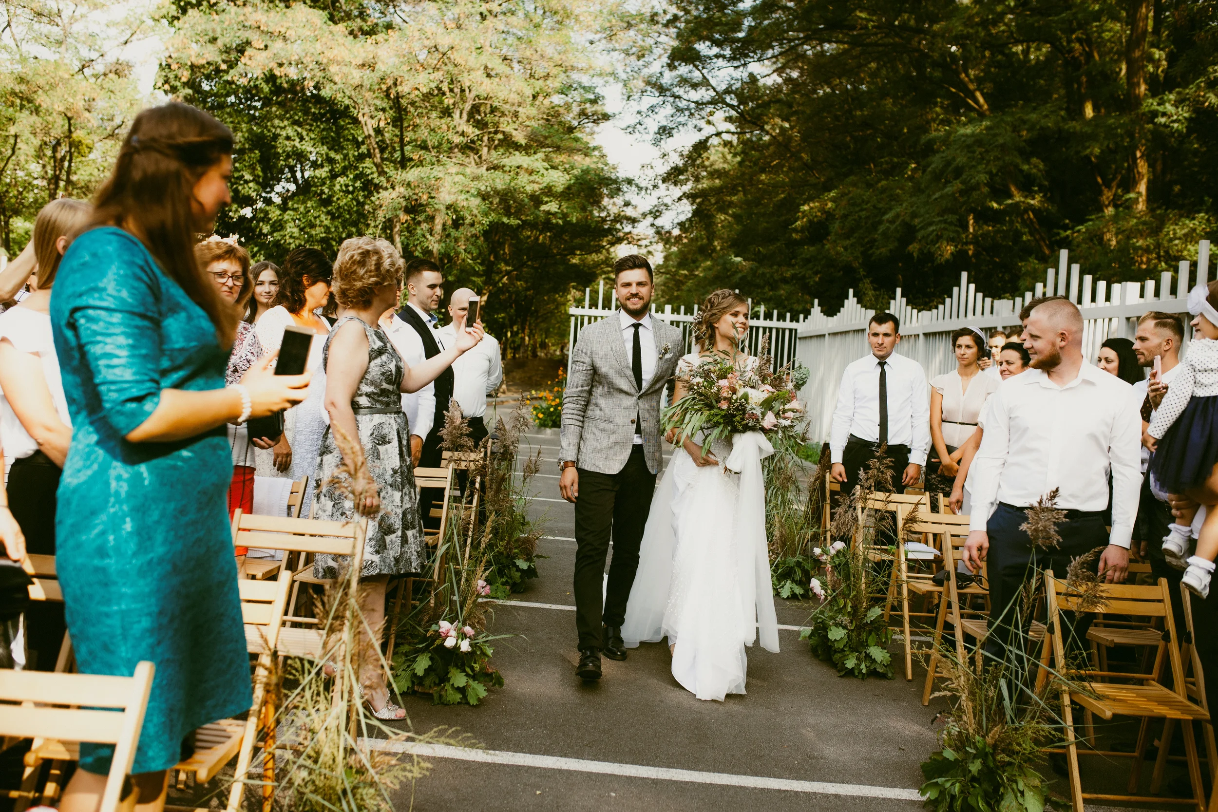 Hochzeitspaar läuft eine Straße entlang, umgeben von Gästen, die Fotos machen, im Freien mit Bäumen im Hintergrund