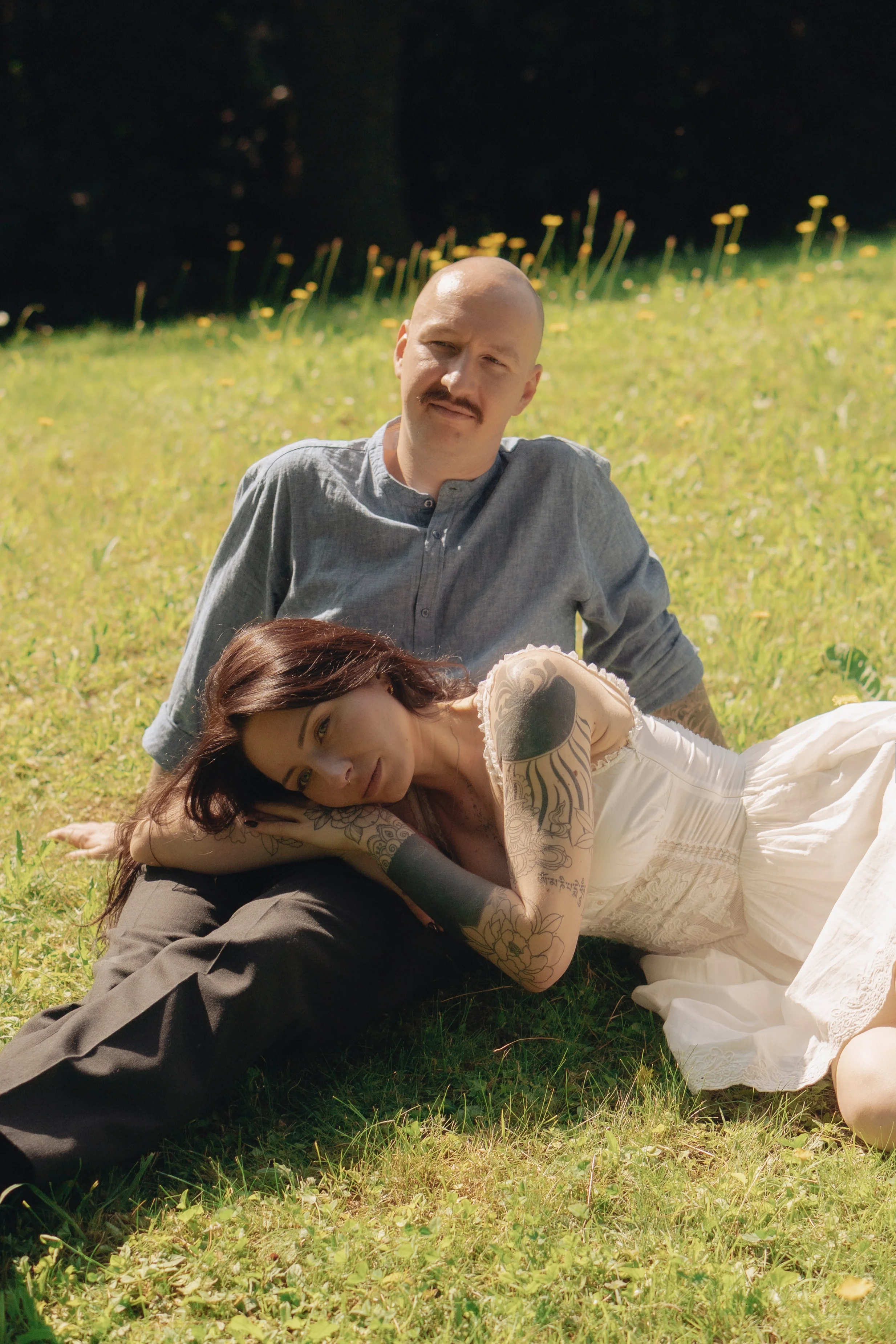 Ein Mann mit Schnurrbart und Glatze sitzt im Gras, während eine Frau mit langen dunklen Haaren und Tattoos auf dem Arm vor ihm liegt, beide in einer Wiese mit gelben Blumen an einem sonnigen Tag.