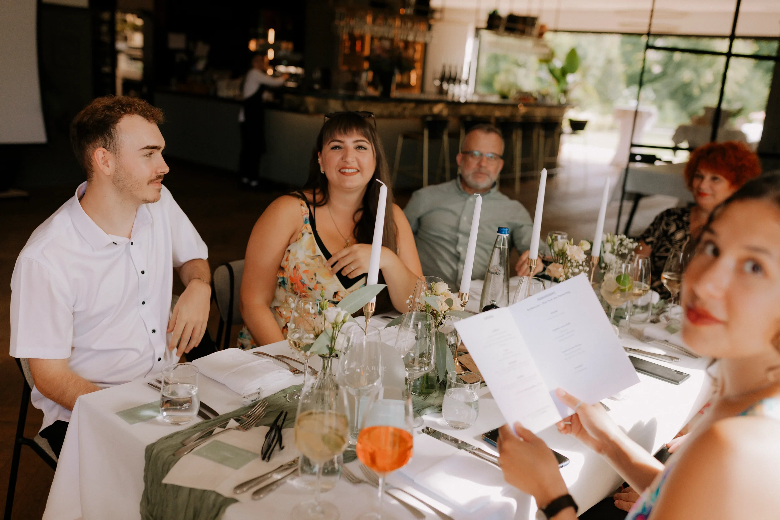 Mehrere Menschen sitzen an einem festlich gedeckten Tisch bei einer Feierlichkeit in einem Restaurant oder einer Eventlocation, mit Dekorationen wie Kerzen und Blumen, während im Hintergrund ein Barbereich sichtbar ist.