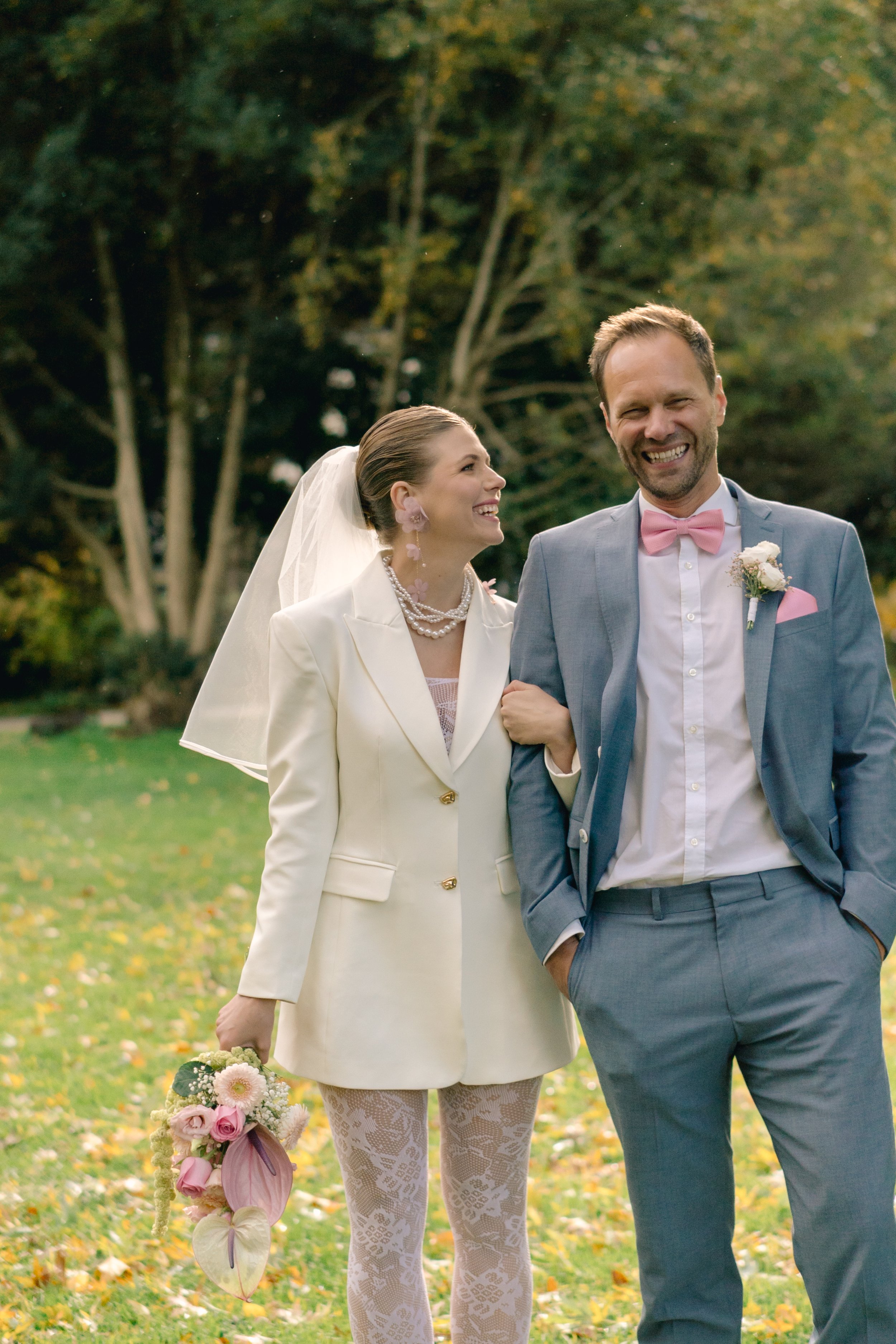 Ein lächelndes Paar bei einer Hochzeit im Freien, die Frau trägt einen weißen Anzug mit Perlen und einen Schleier, der Mann trägt einen grauen Anzug mit rosa Fliege und Ansteckblume, im Hintergrund Bäume und Blätter auf dem Boden.