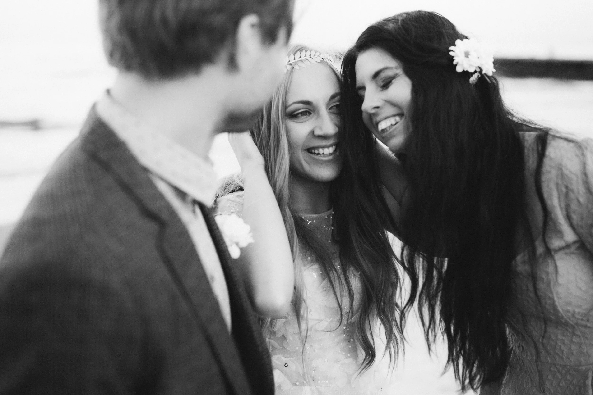 Zwei Frauen lachen eng beieinander, eine trägt eine Blumenkette im Haar, die andere hat eine große Blume im Haar. Ein Mann ist leicht unscharf im Vordergrund sichtbar.