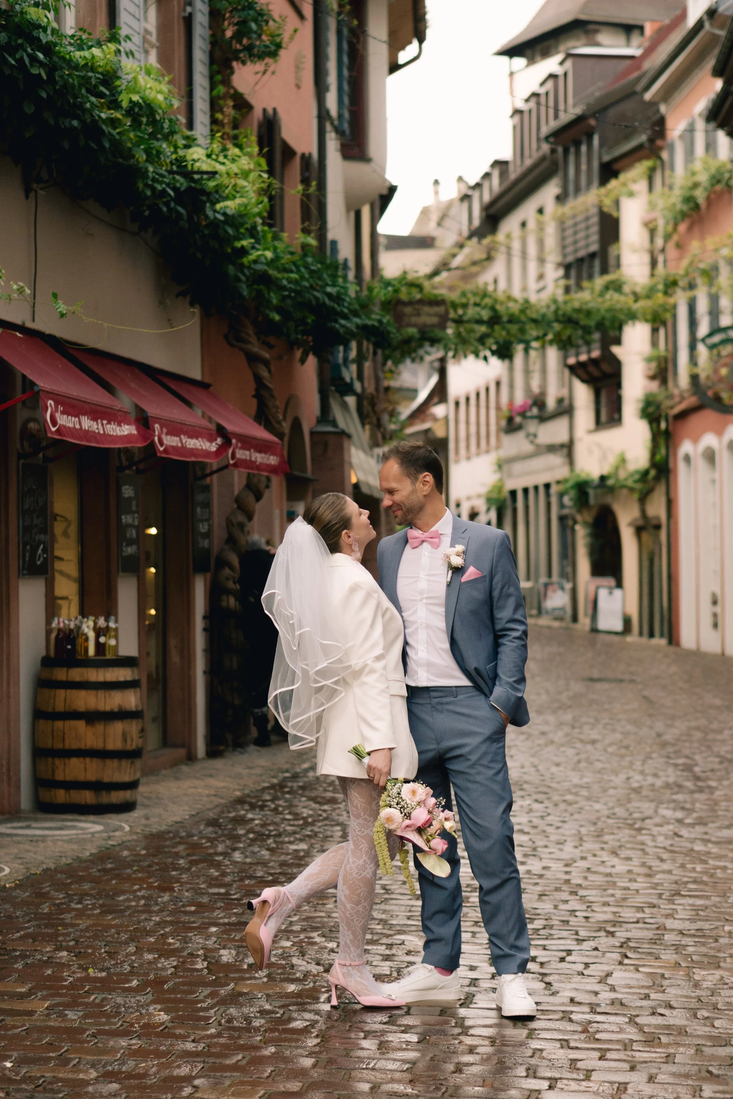Ein Paar bei einer Hochzeit auf einer kleinen europäischen Strae, das sich liebevoll ansieht, mit Kopf an Kopf und lachend. Die Frau trägt ein weis Hochzeitskleid und die hohe Hackenschuhe, der Mann einen Anzug mit rosa Fliege. Sie halten einen Blu