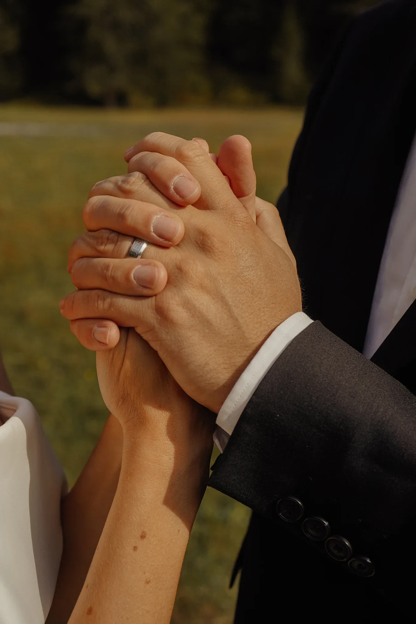 Nahaufnahme von zwei Personen, die sich gegenseitig die Hände halten bei einer Hochzeit im Freien. Eine Person trägt einen schwarzen Anzug und die andere eine weiße Kleidung.