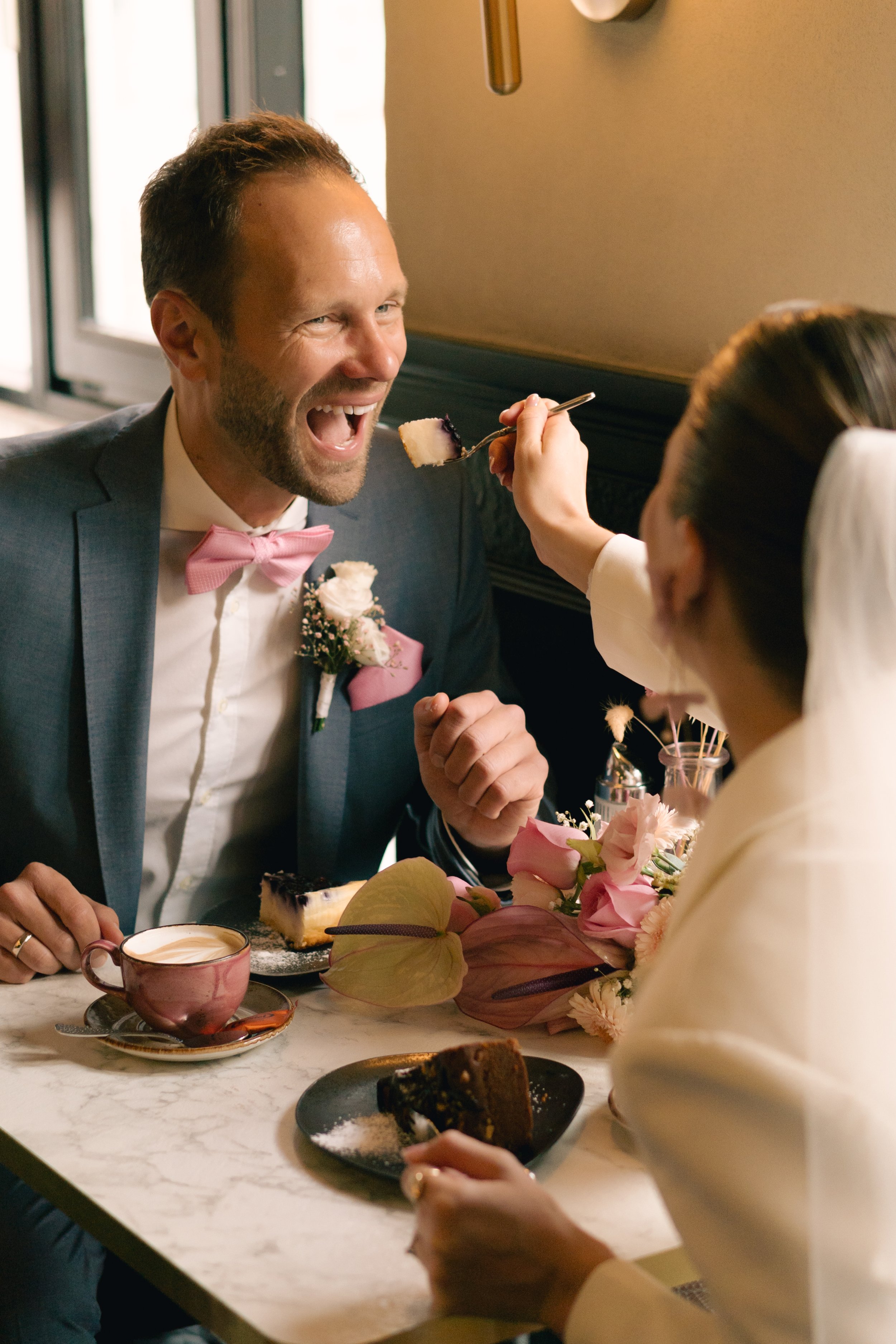 Ein Mann in formeller Kleidung, mit rosa Fliege und Blumen im Revers, lacht, während eine andere Person ihm ein Stück Kuchen auf die Gabel gibt. Es gibt Blumen und Tassen auf einem Tisch. Es scheint eine festliche Szene zu sein.