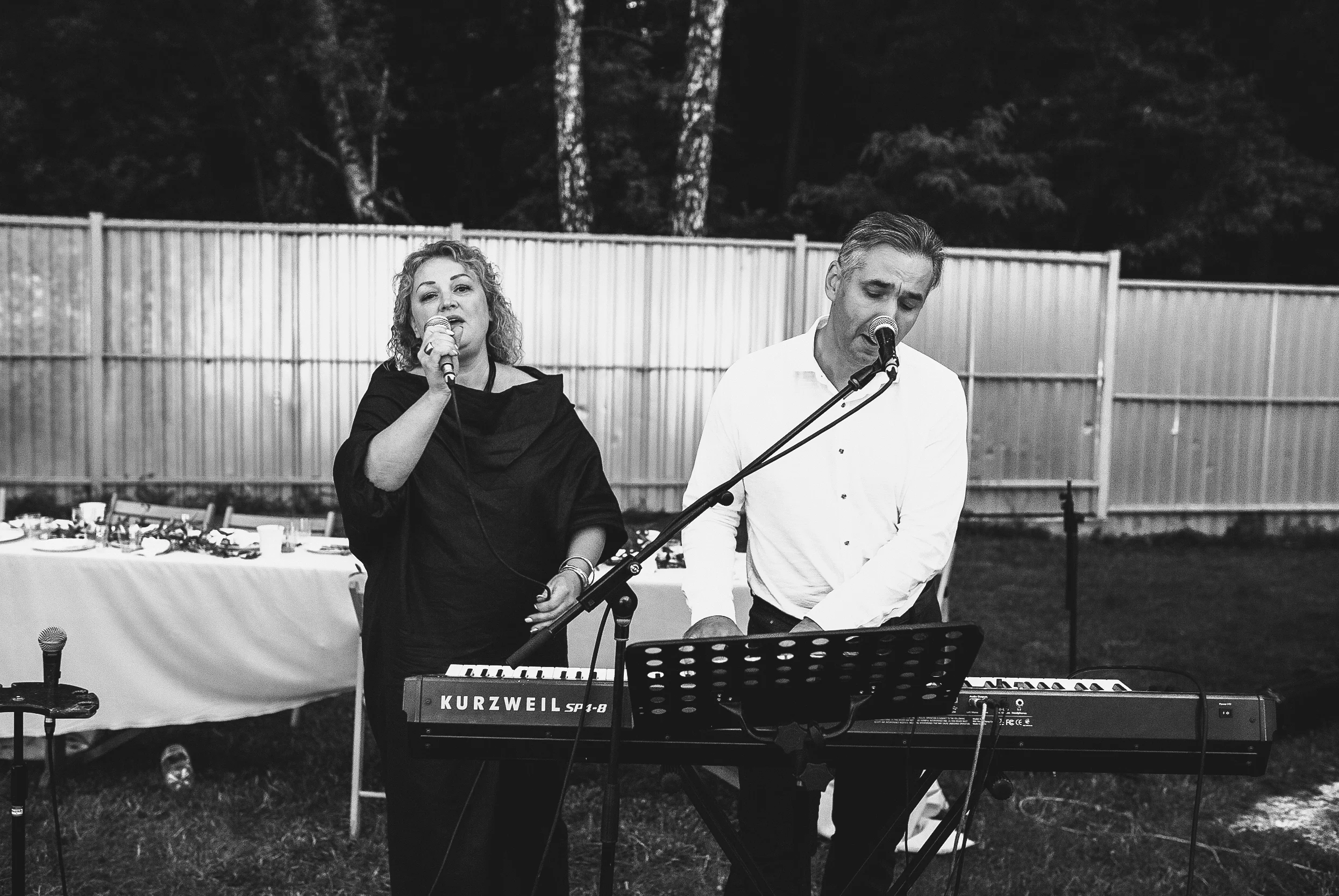 Zwei Musiker, eine Frau singt in ein Mikrofon, ein Mann spielt Keyboard bei einer Outdoor-Veranstaltung, im Hintergrund ein Holzzaun und Bäume, schwarz-weiß