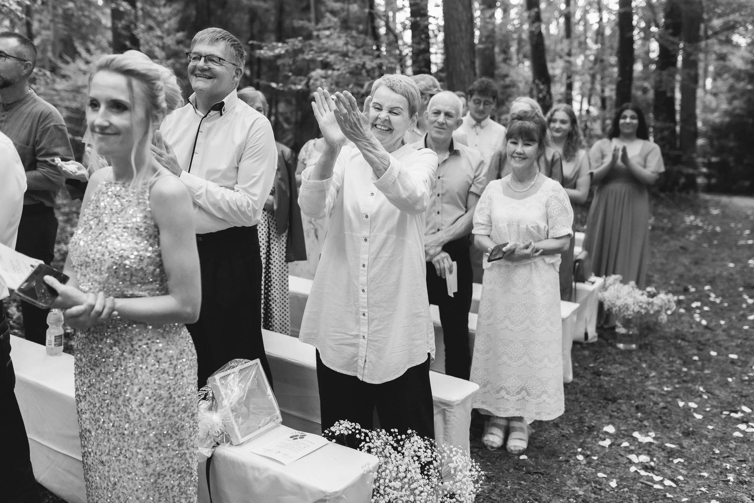 Eine Gruppe von Menschen, die bei einem Außenereignis im Wald lachen und sich freuen, einige klatschen. Im Vordergrund ist eine Frau in einem glitzernden Kleid und eine andere Frau, die einen Tisch mit Dekorationen und Blumen hat.