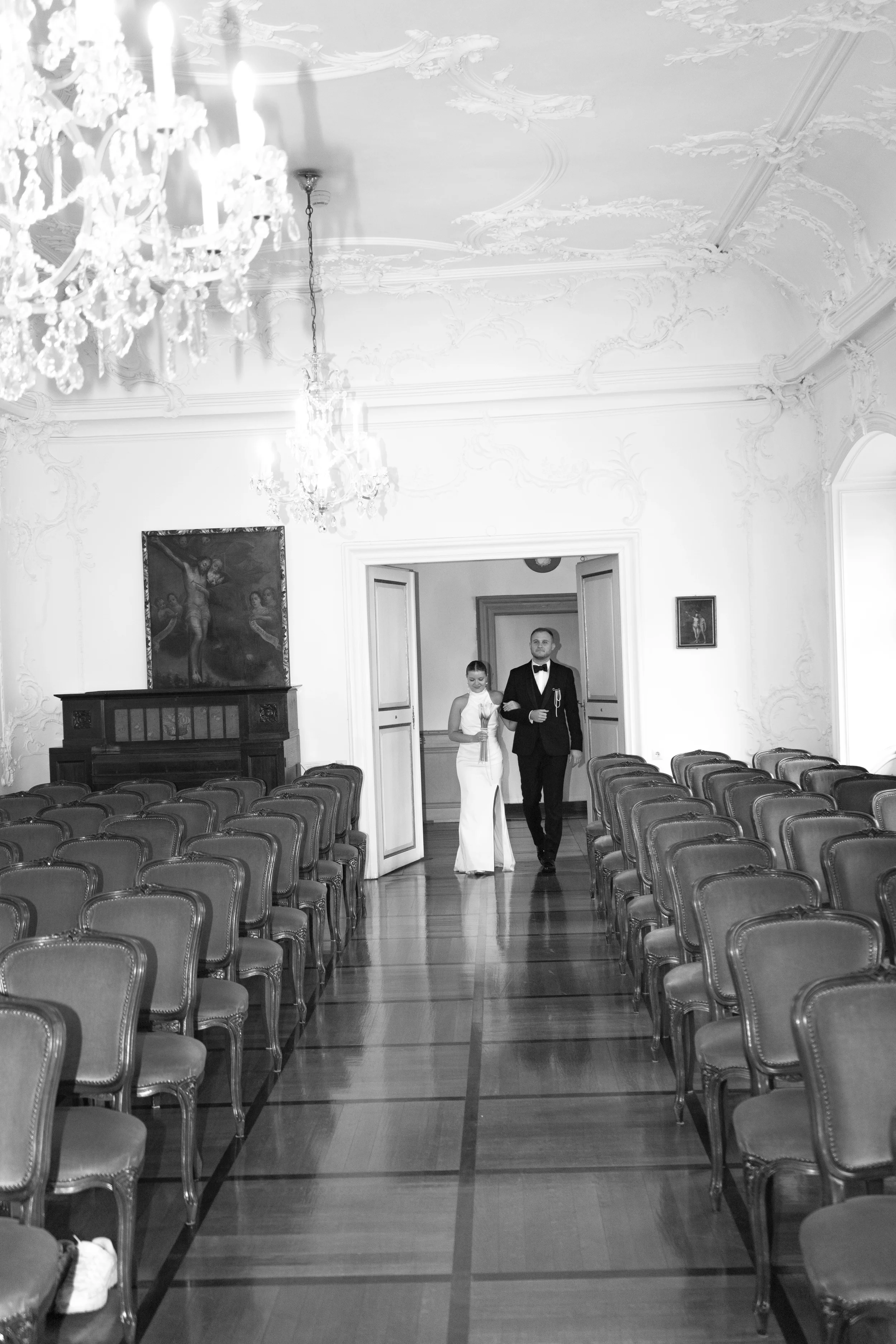 Ein eleganter Saal mit hohen Decken, ornamentierten Wänden und Kronleuchtern. Eine Frau in einem weißen Kleid und ein Mann in einem schwarzen Anzug gehen durch die Tür in den Saal.