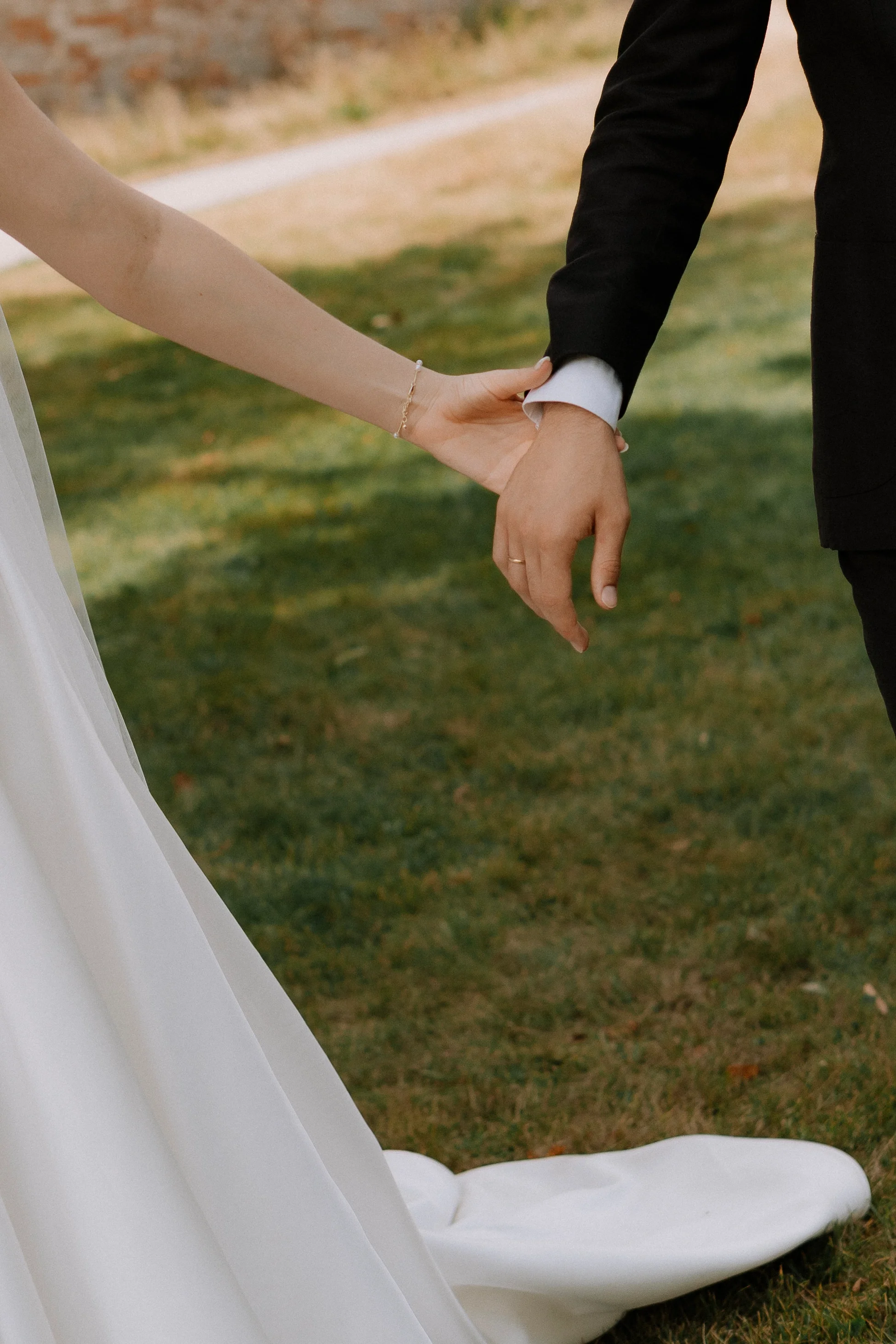 Hände eines Brautpaars, die sich bei einer Hochzeit auf einer Wiese halten, mit einem weißen Brautkleid und einem schwarzen Anzug.