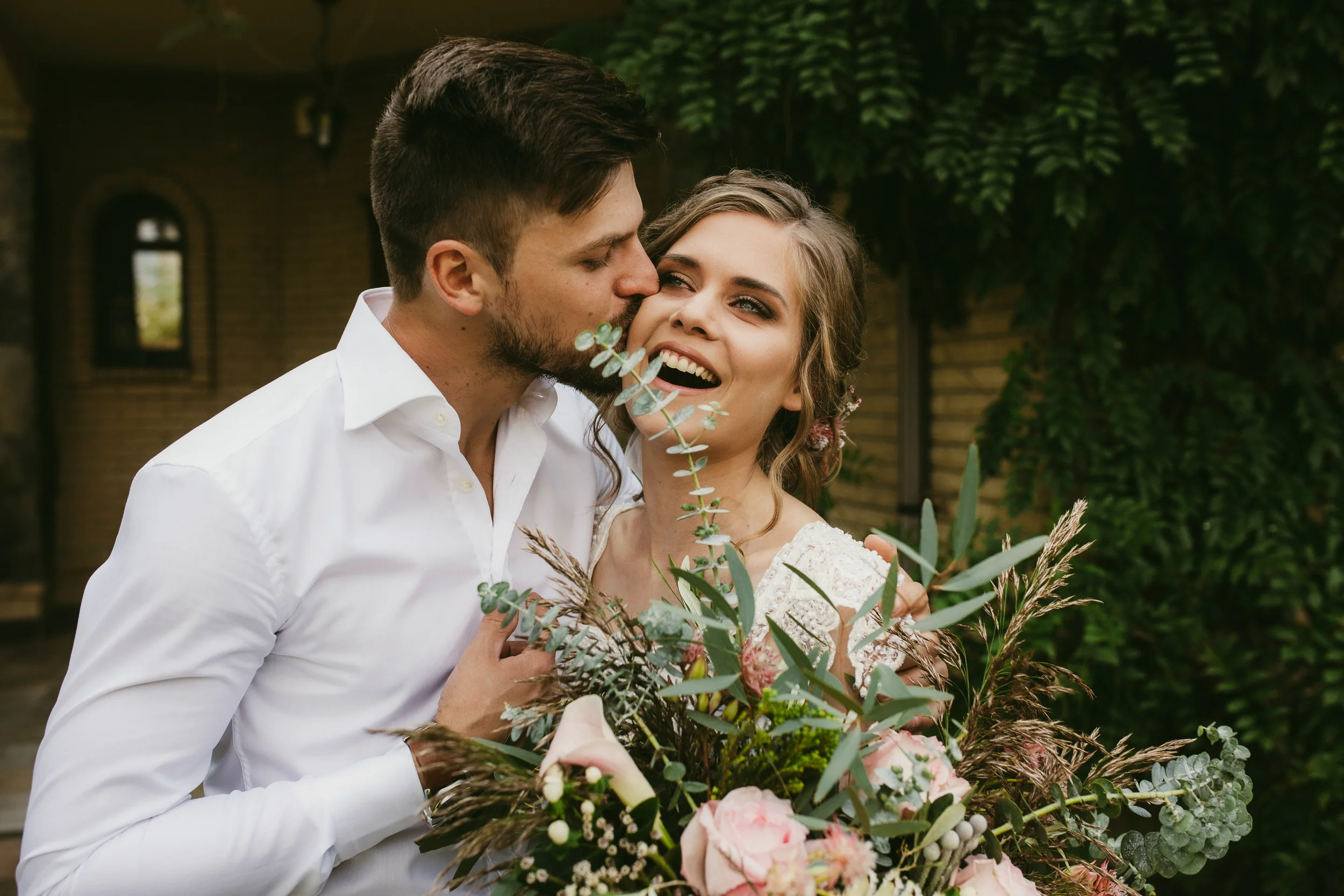 Ein glückliches Paar bei einer Hochzeit, das die Braut liebevoll küsst, im Garten mit grünem Laub im Hintergrund.