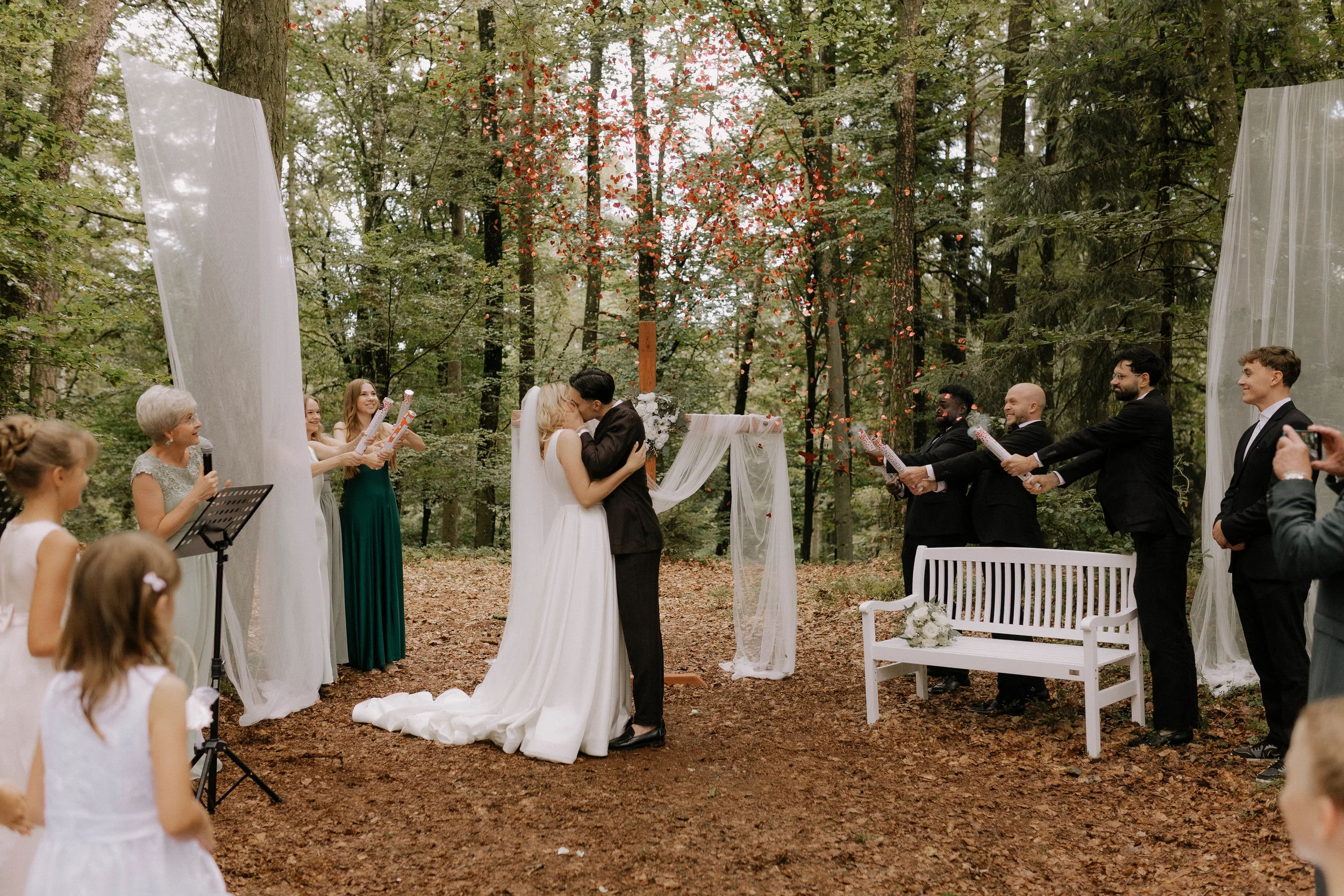 Ein Hochzeitsritual im Wald, bei dem das Brautpaar sich küsst, während die Gäste zuschauen. Es ist ein festliches Ereignis mit einer naturverbundenen Atmosphäre, inklusive Blumendekoration und einer achteckigen Sitzbank für das Brautpaar.