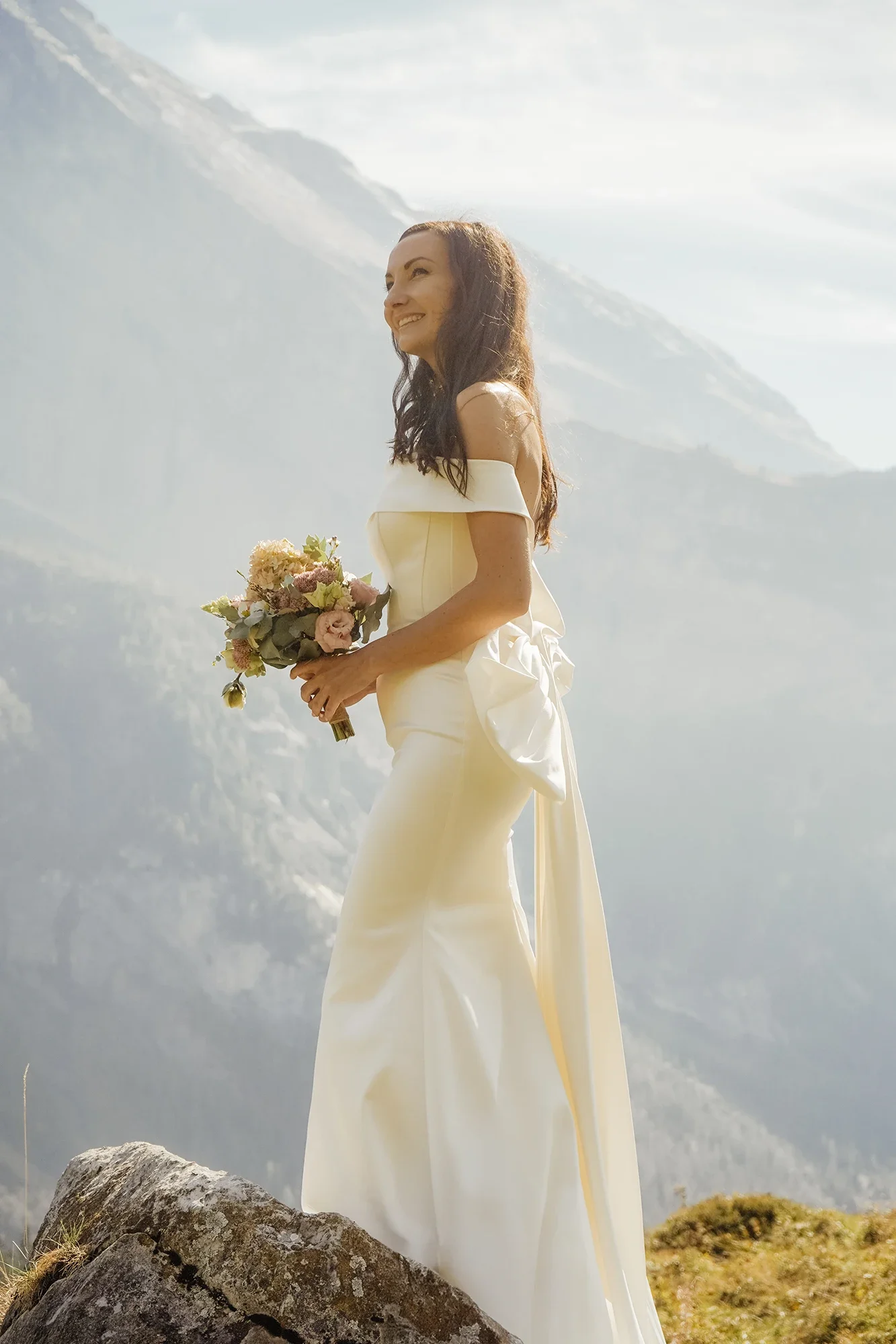 Eine lachende Frau in einem weißen Kleid hält einen Blumenstrauß vor einer bergigen Landschaft mit Schnee bedeckten Gipfeln.