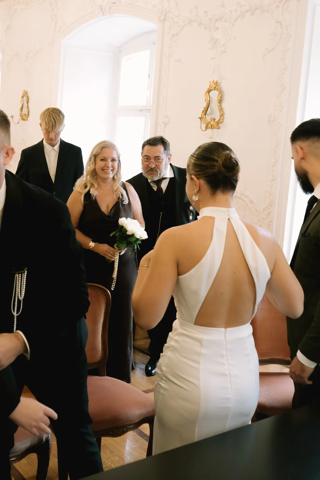 Eine Hochzeitsszene mit Braut in weißem Kleid und Gästen in formeller Kleidung in einem eleganten Raum.